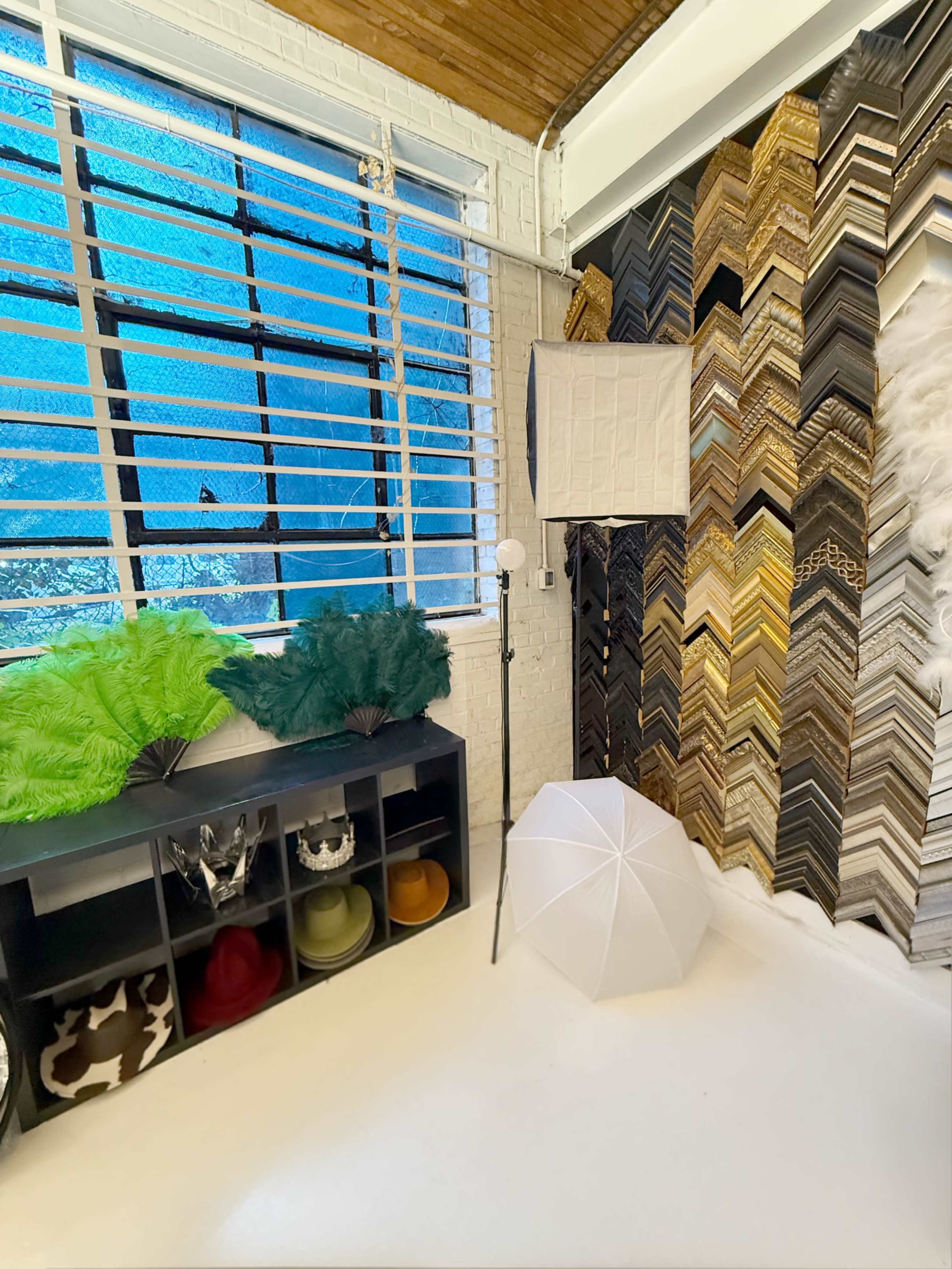 The image shows a brightly lit room featuring a wall with a chevron pattern of picture frames, a white umbrella, decorative greenery, and shelves containing various hats and masks.