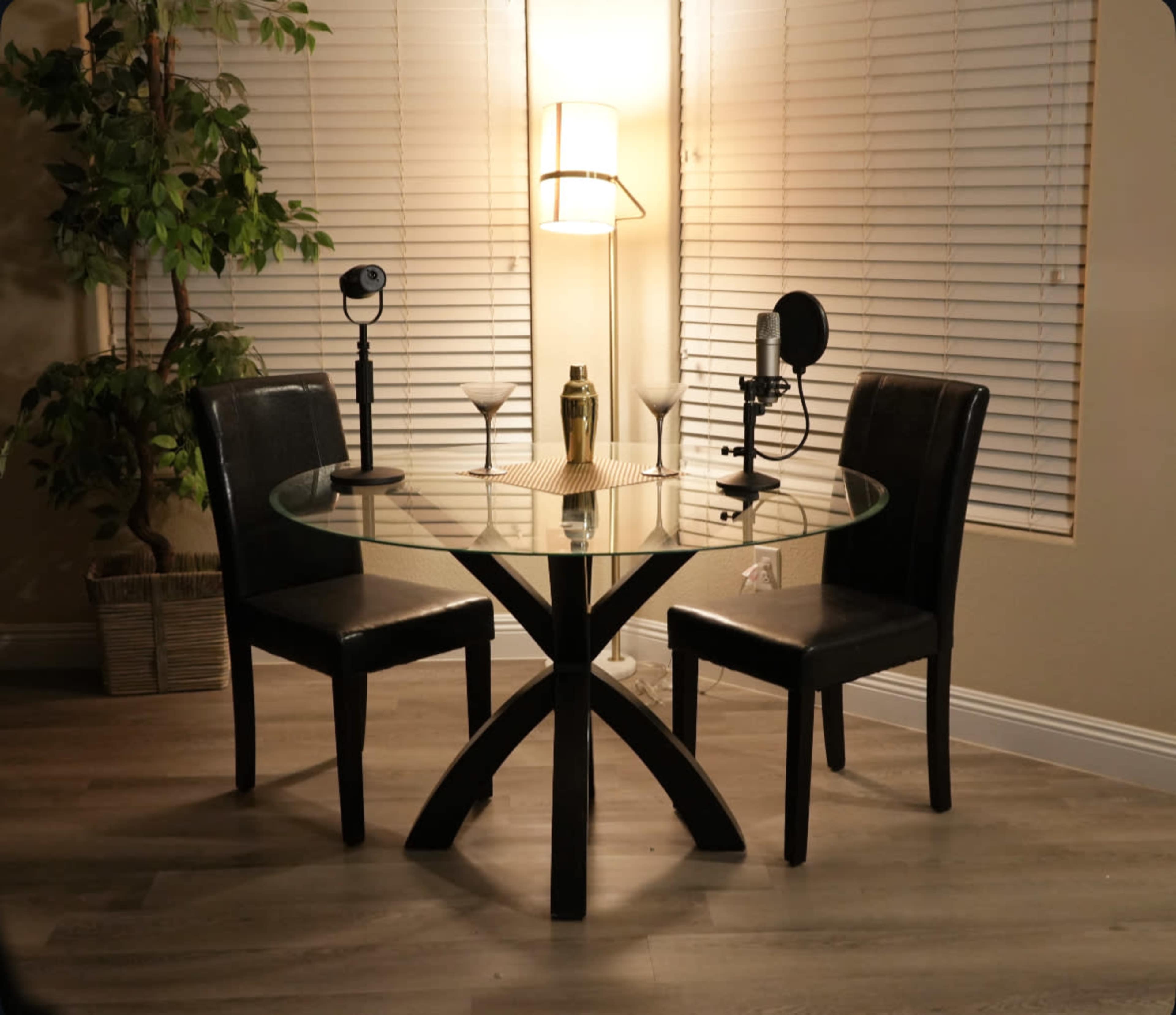 A round glass table with two black chairs is set up in a well-lit room, featuring a lamp and audio recording equipment on the table.