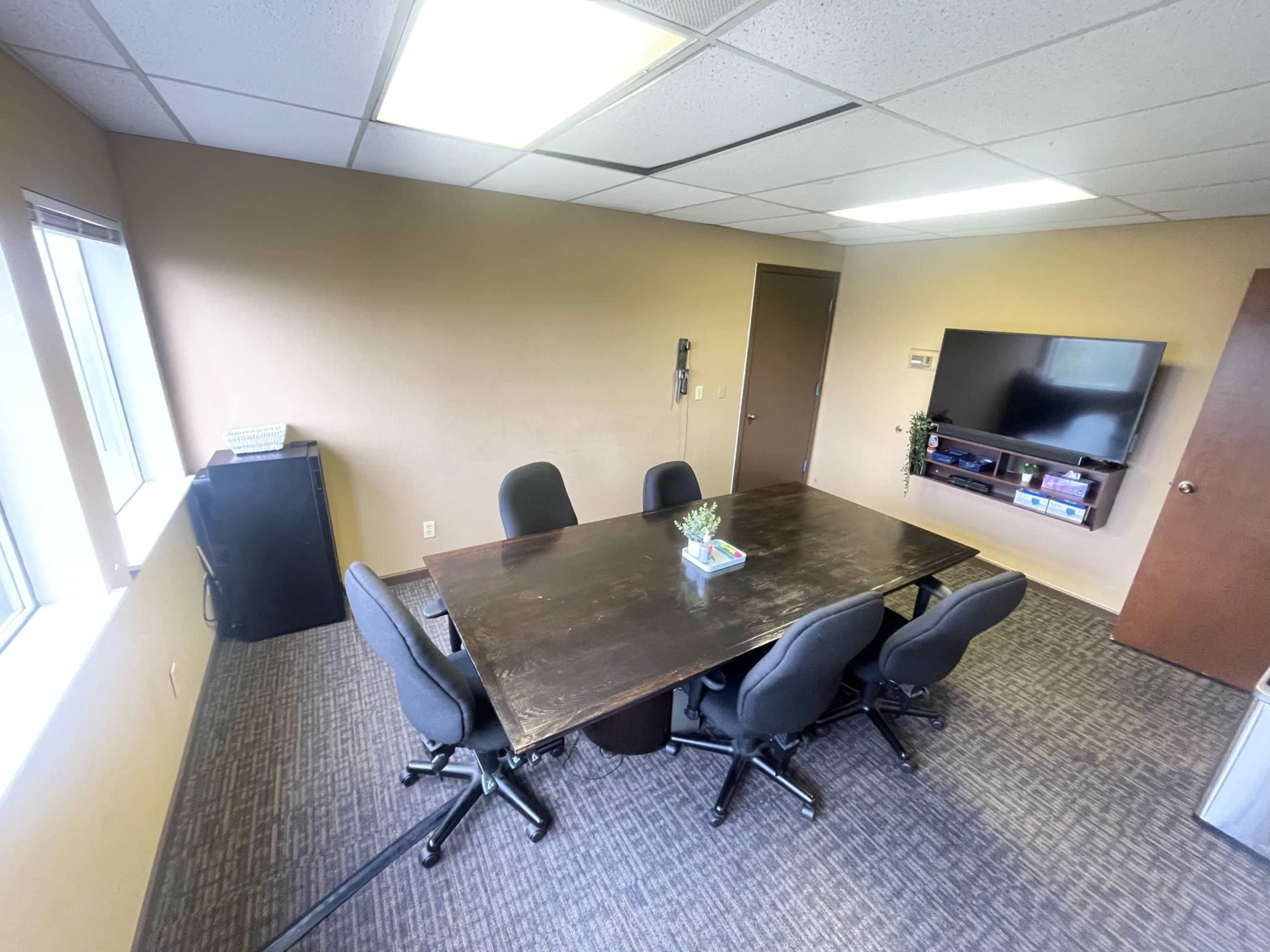 A conference room with a large table surrounded by black chairs, a television mounted on the wall, and two windows allowing natural light.