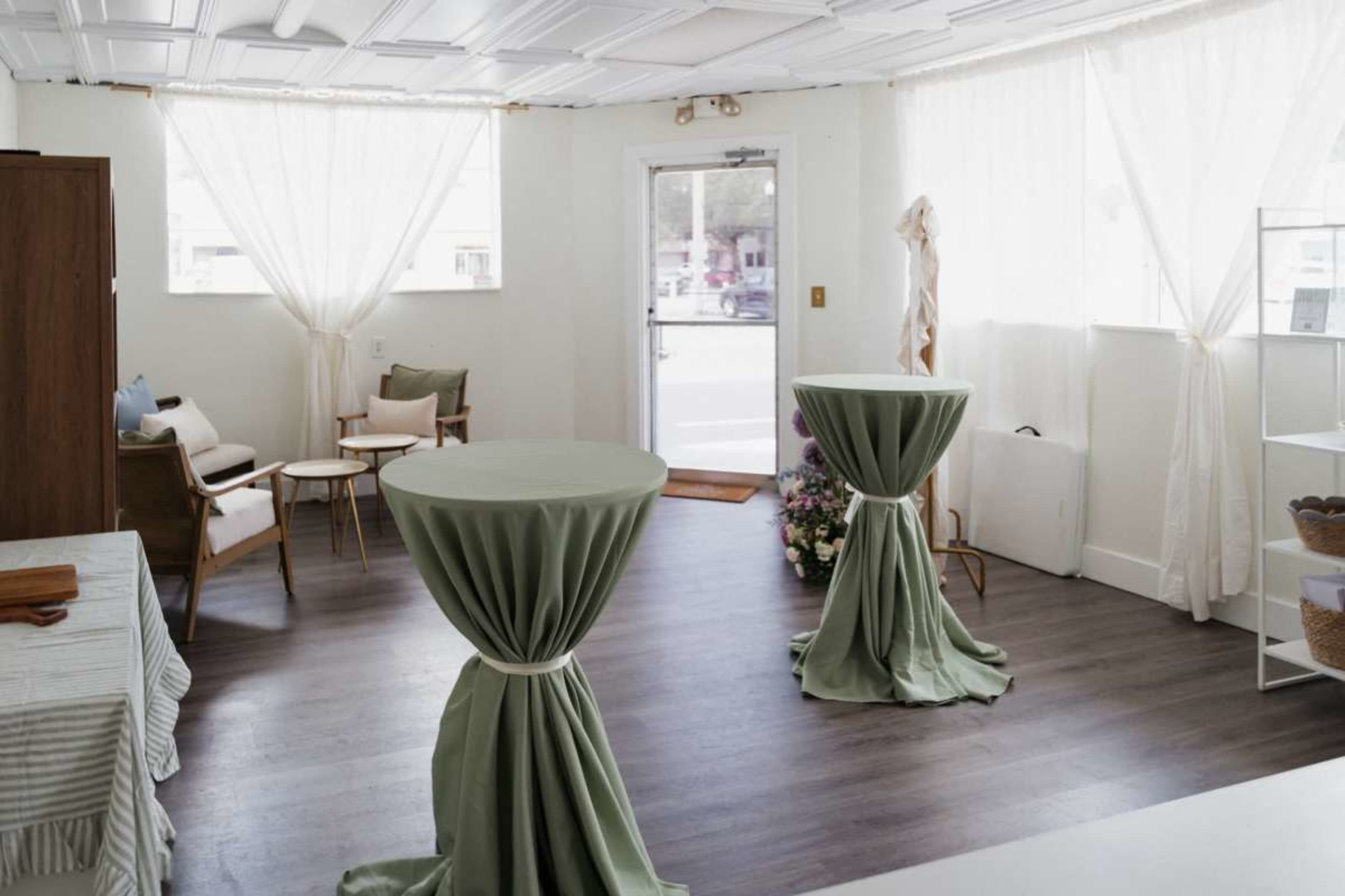 The image shows a bright, uncluttered room featuring two tall, round tables covered with green cloths, and a seating area with chairs near a large window.