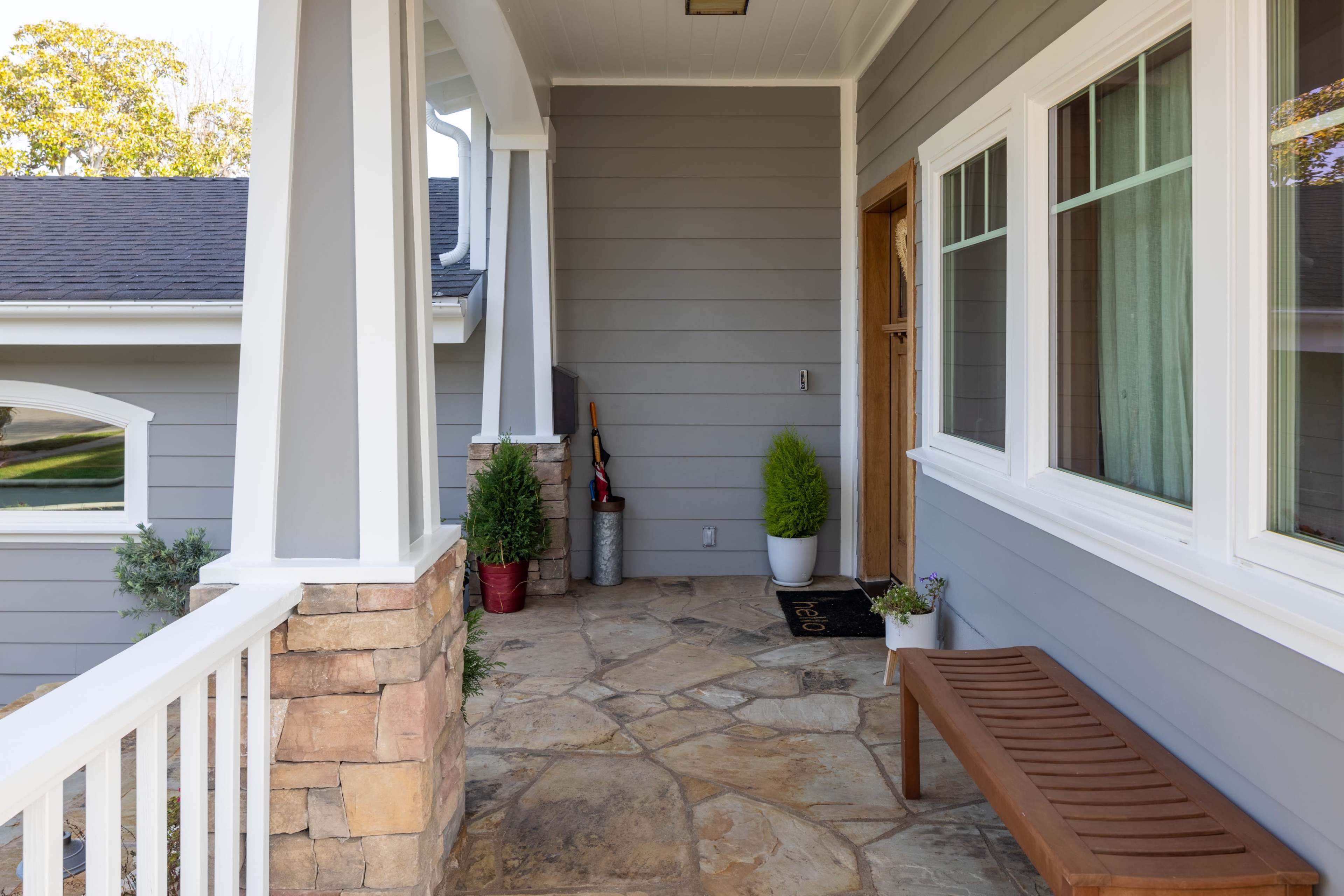 A front porch with stone flooring, a wooden bench, and potted plants beside a door with large windows.