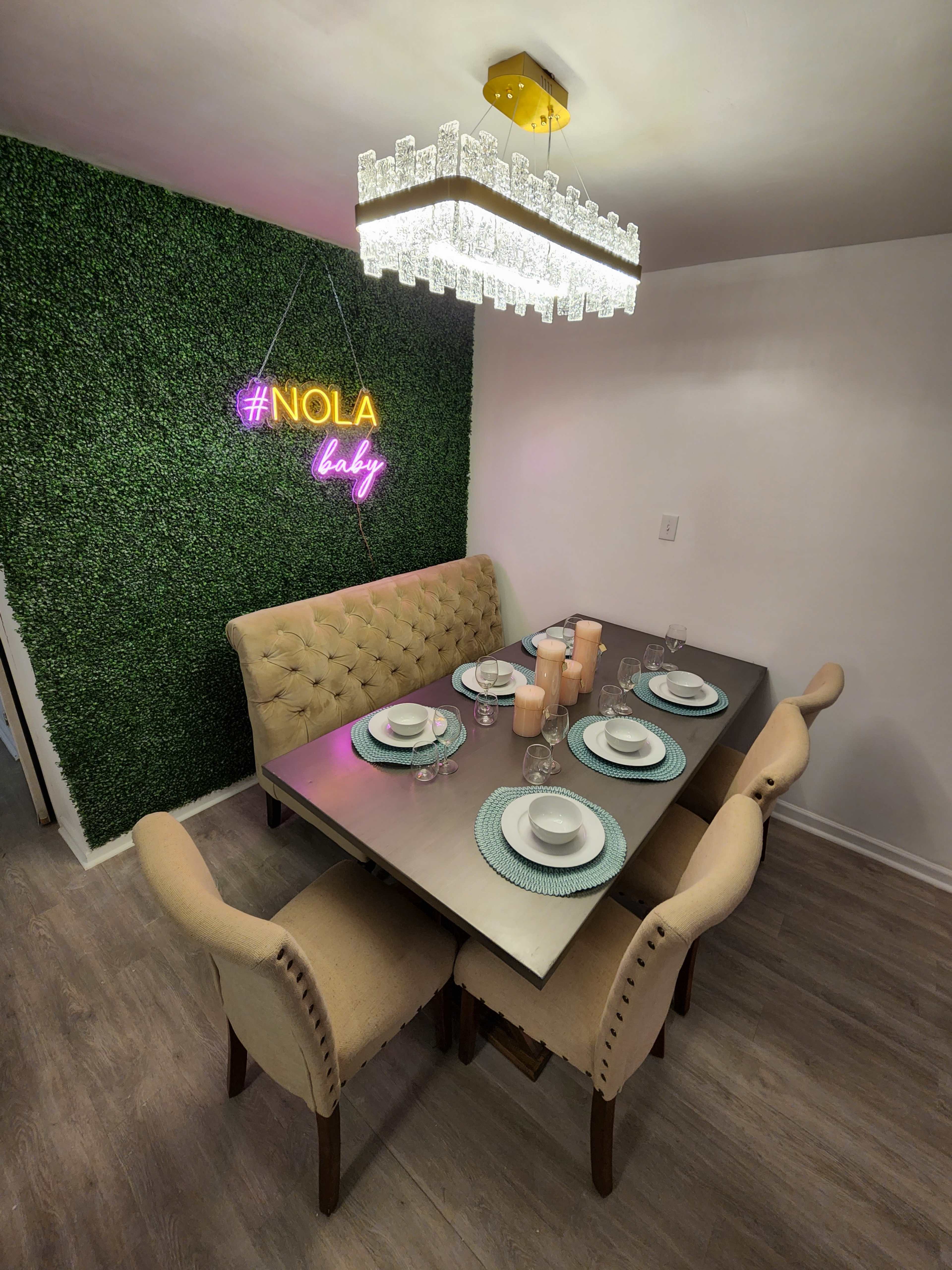 A dining area with a rectangular table set for six, featuring a green wall adorned with a neon sign that reads "#NOLA baby" and a modern chandelier hanging above.