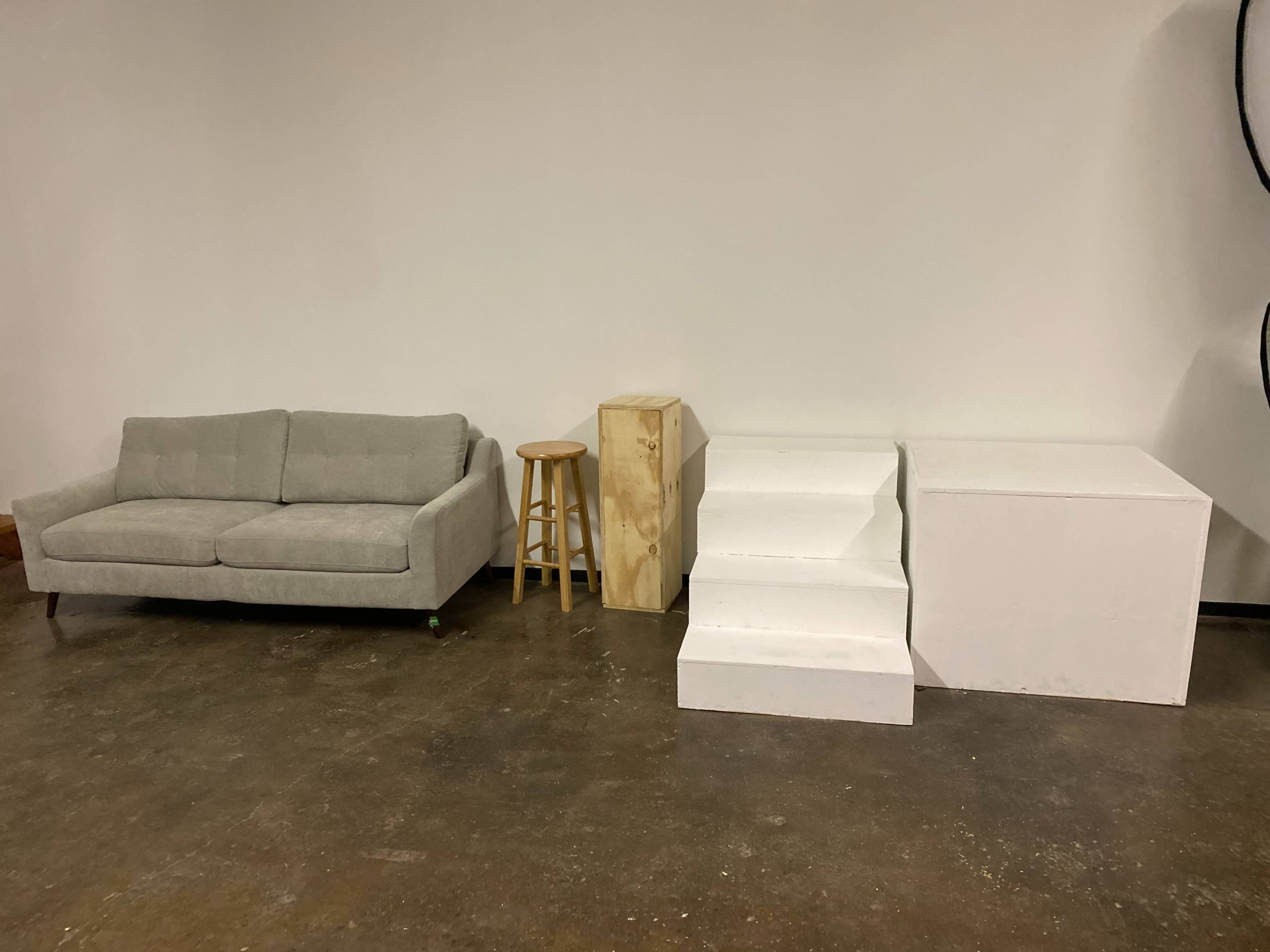 A light gray couch, a wooden stool, a wooden block, and a white staircase structure arranged on a concrete floor against a plain wall.