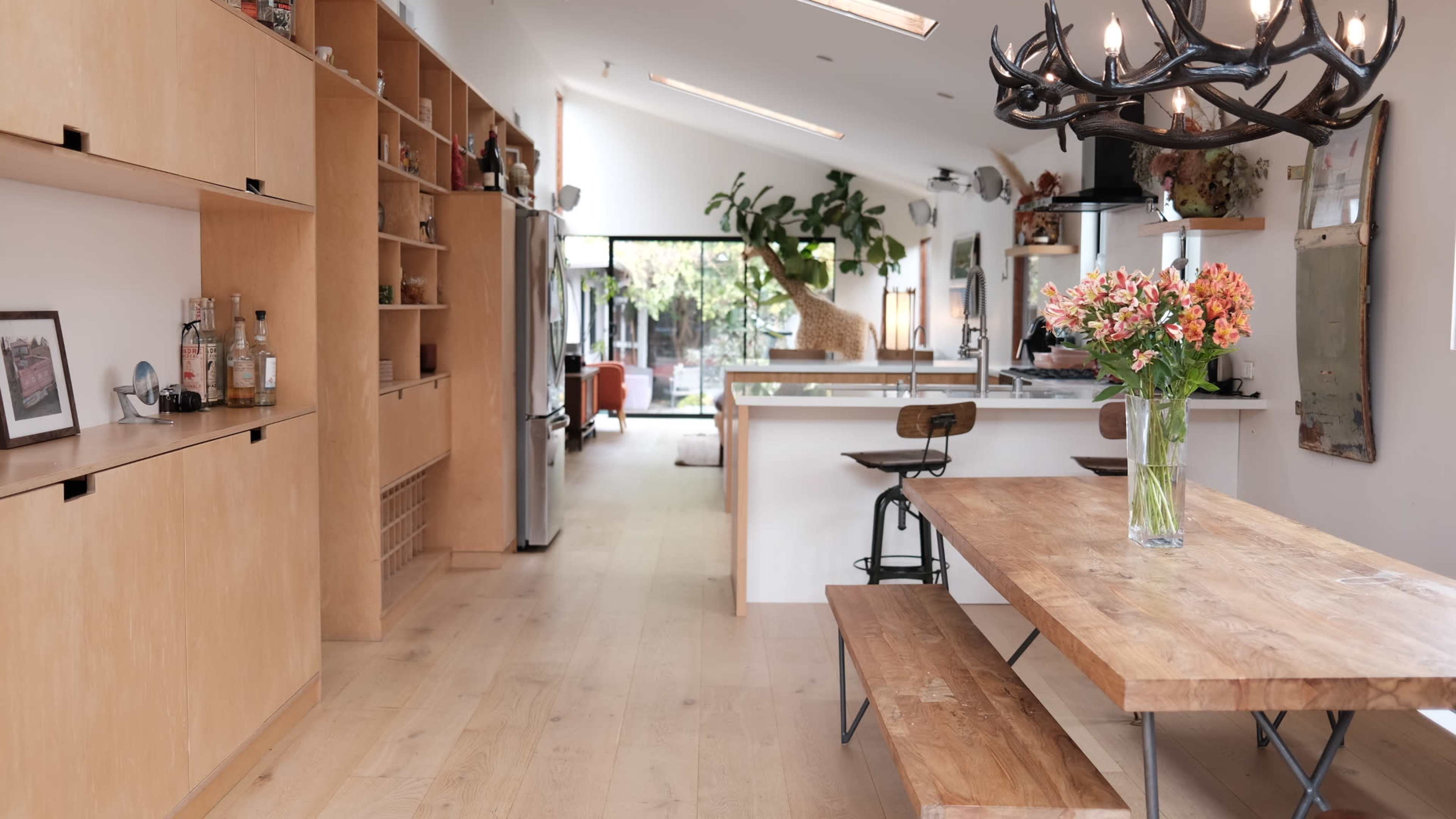 A modern kitchen with wooden cabinetry, a central dining table, and large windows that open to a living area filled with plants and natural light.