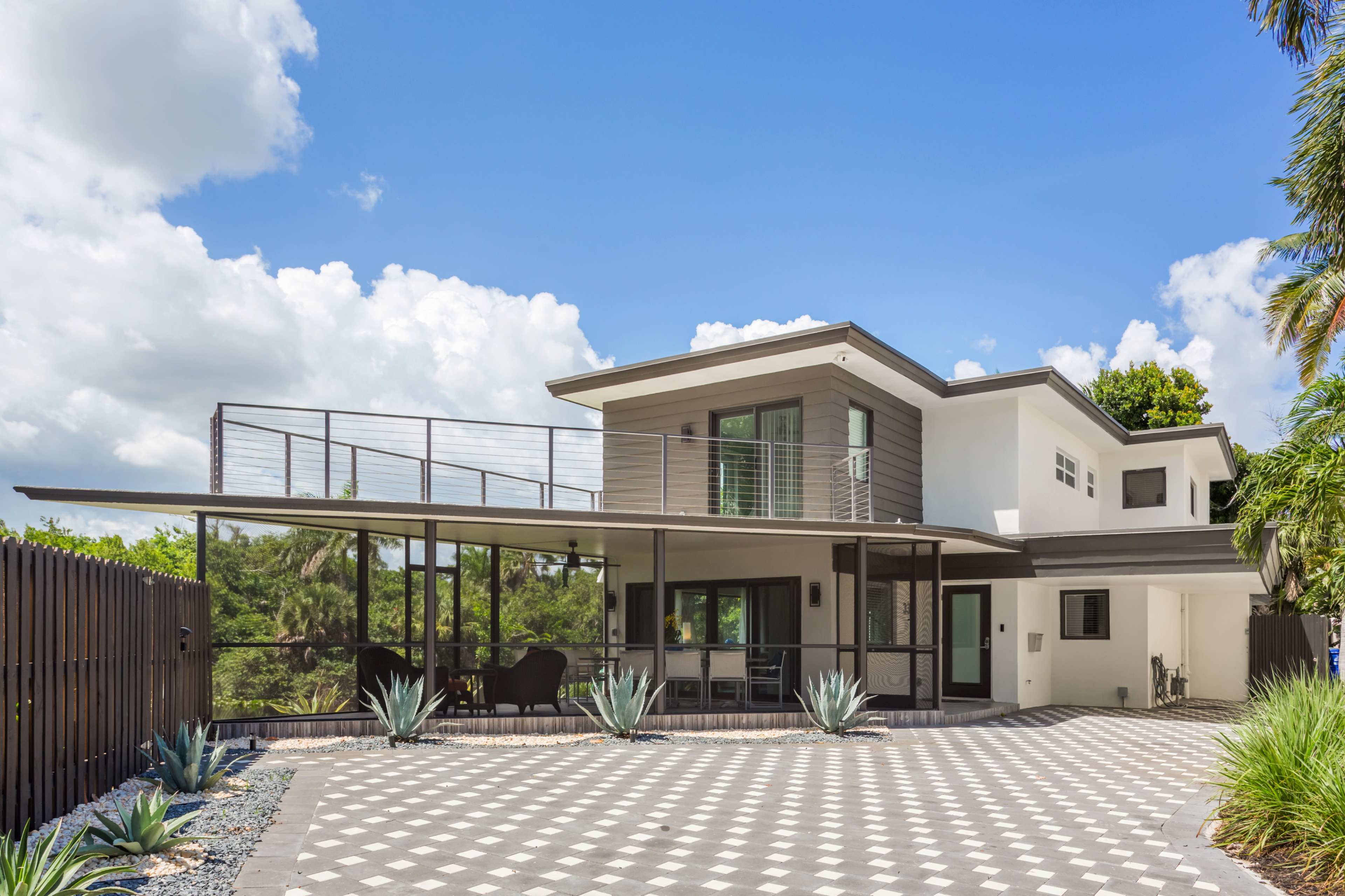 The image shows a modern two-story house with a large balcony and a patterned driveway surrounded by desert plants.