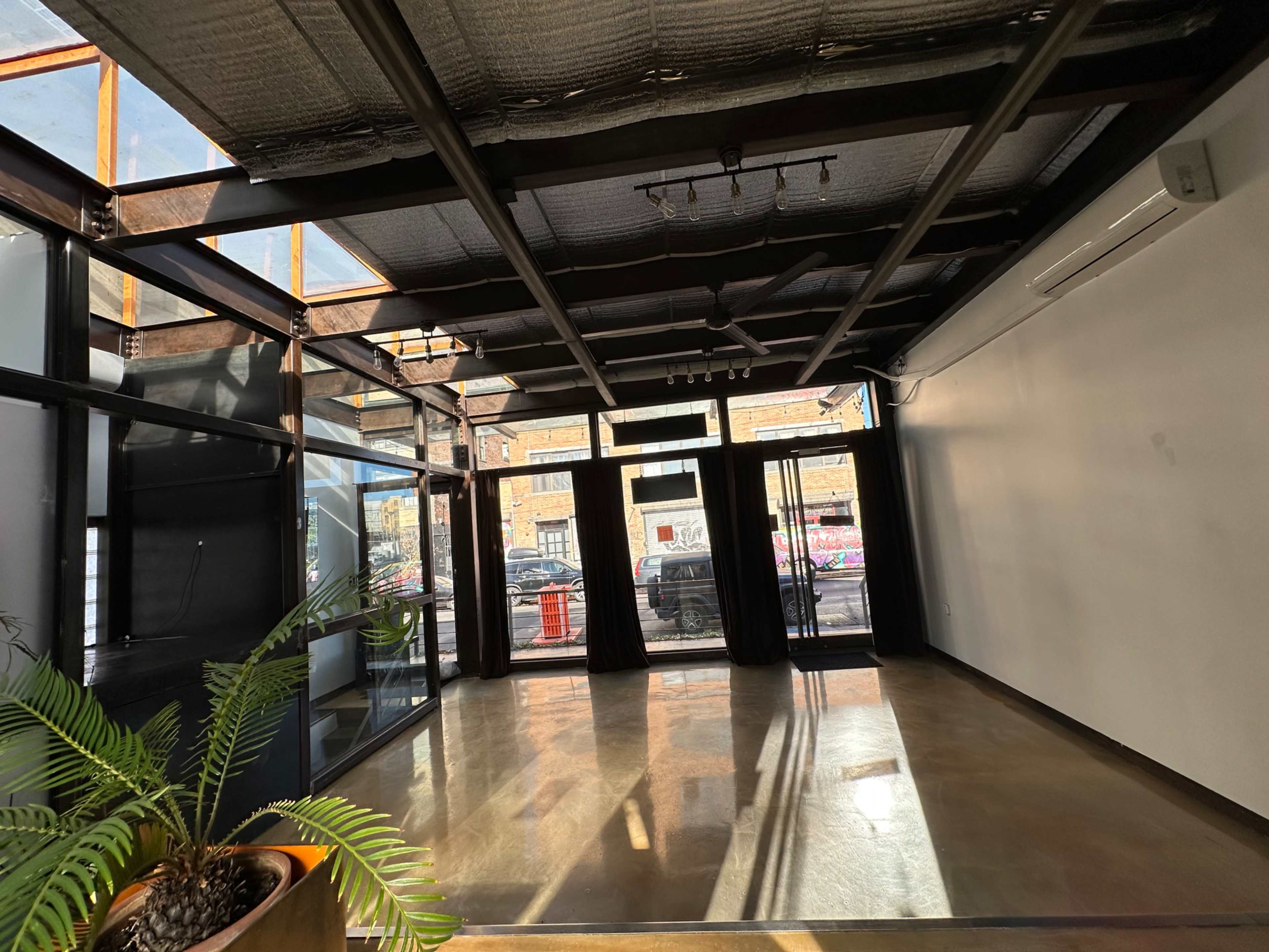 The image shows an empty room with large glass doors and windows, featuring a polished concrete floor and a potted plant in the corner.