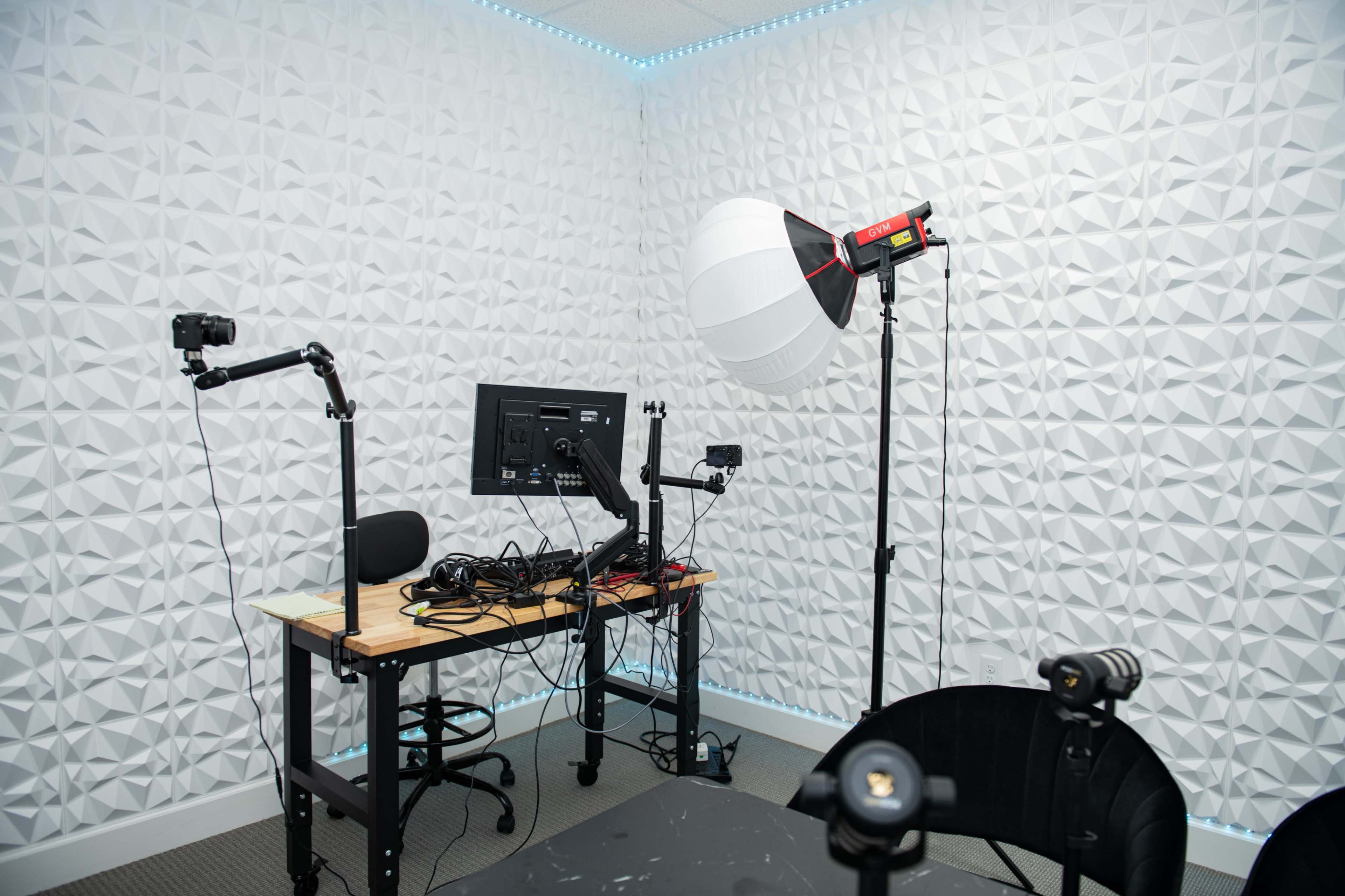 The image shows a small studio setup with a table, multiple cameras mounted on arms, a large softbox light, and a computer monitor, all against a textured white wall.