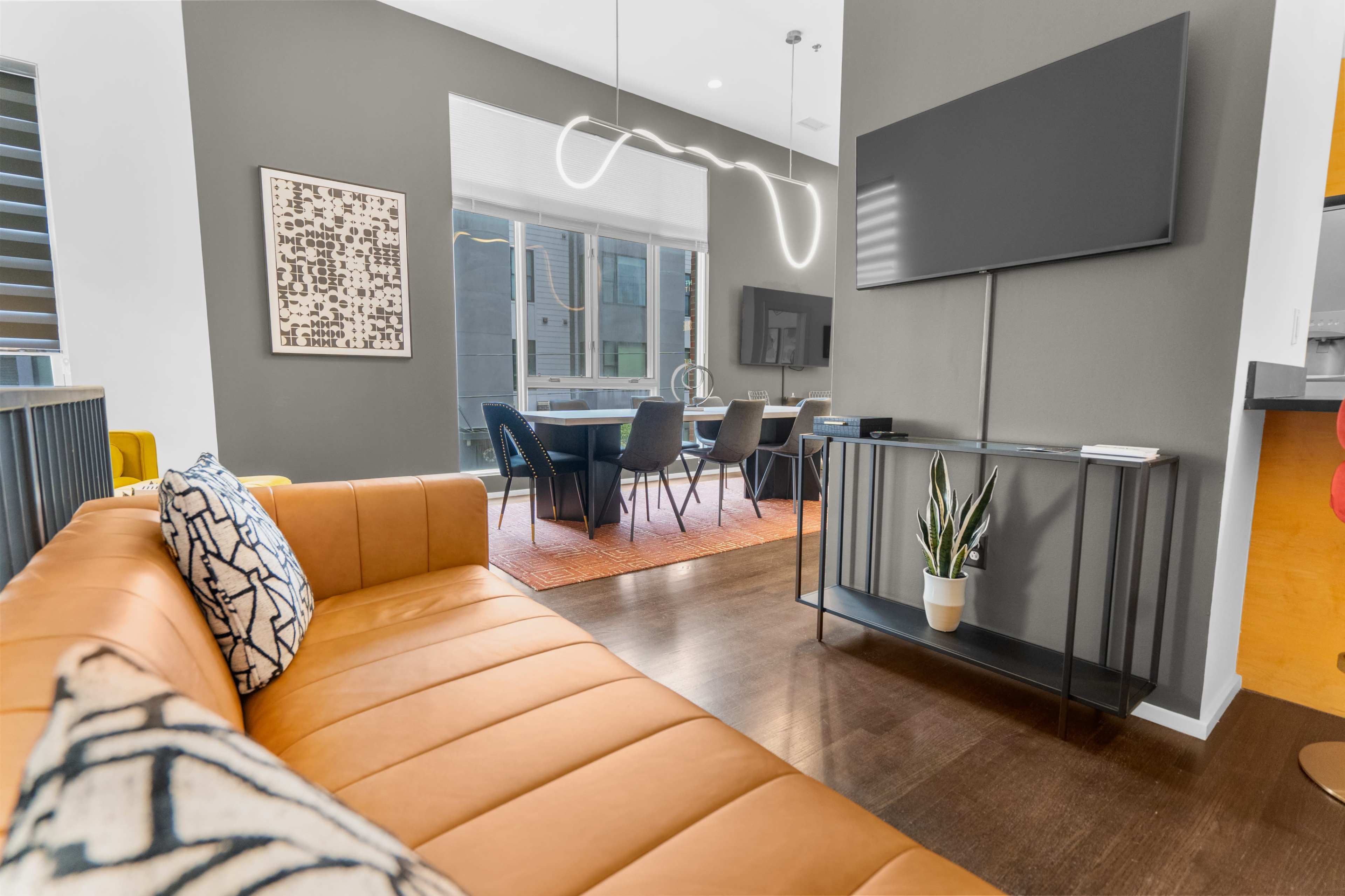 A modern living space features a brown leather sofa, a television mounted on the wall, and a dining area with a table and chairs visible in the background.