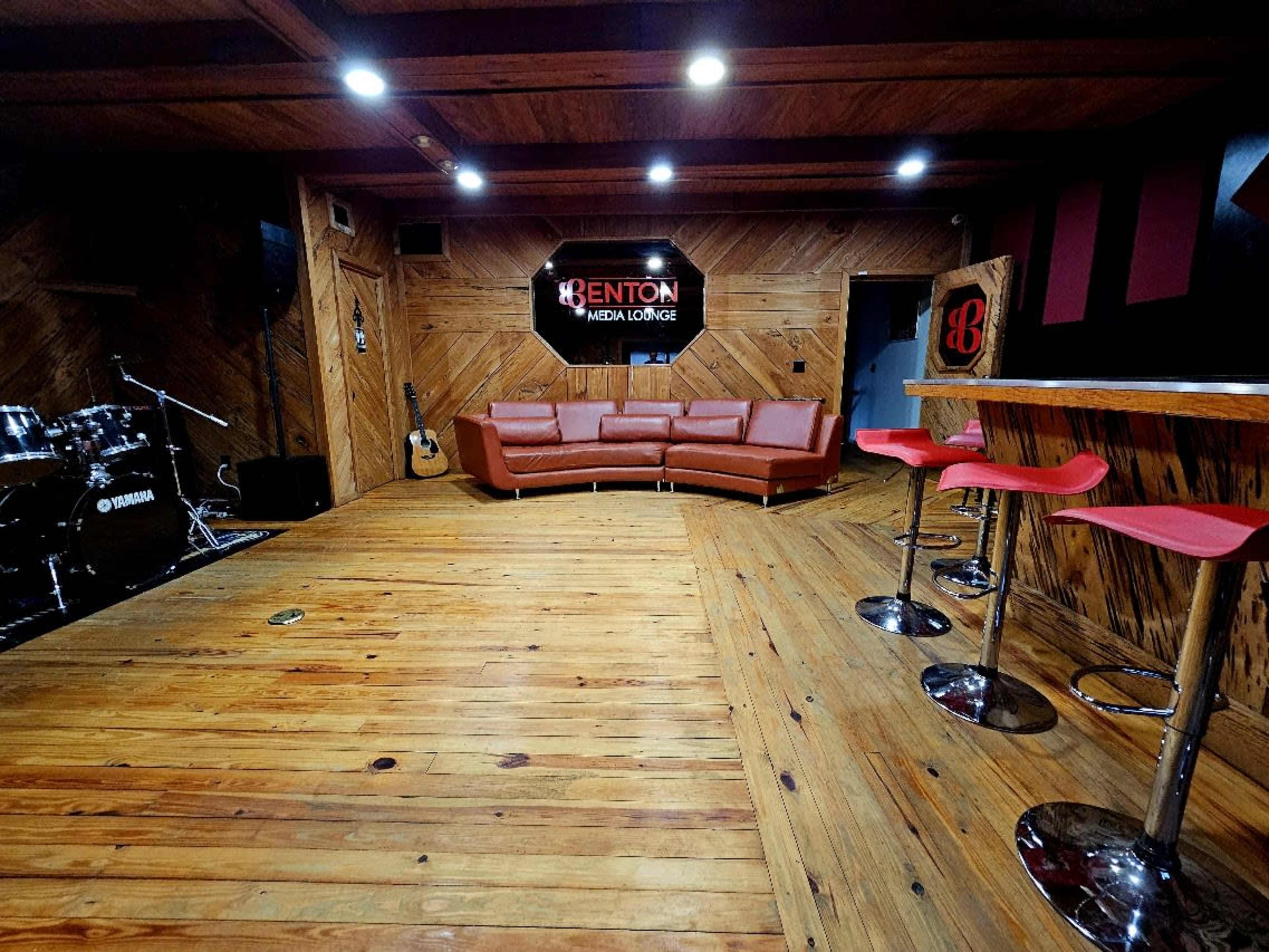 The image shows a cozy media lounge featuring wooden walls, a red leather couch, a bar with high stools, and musical instruments in the corner.