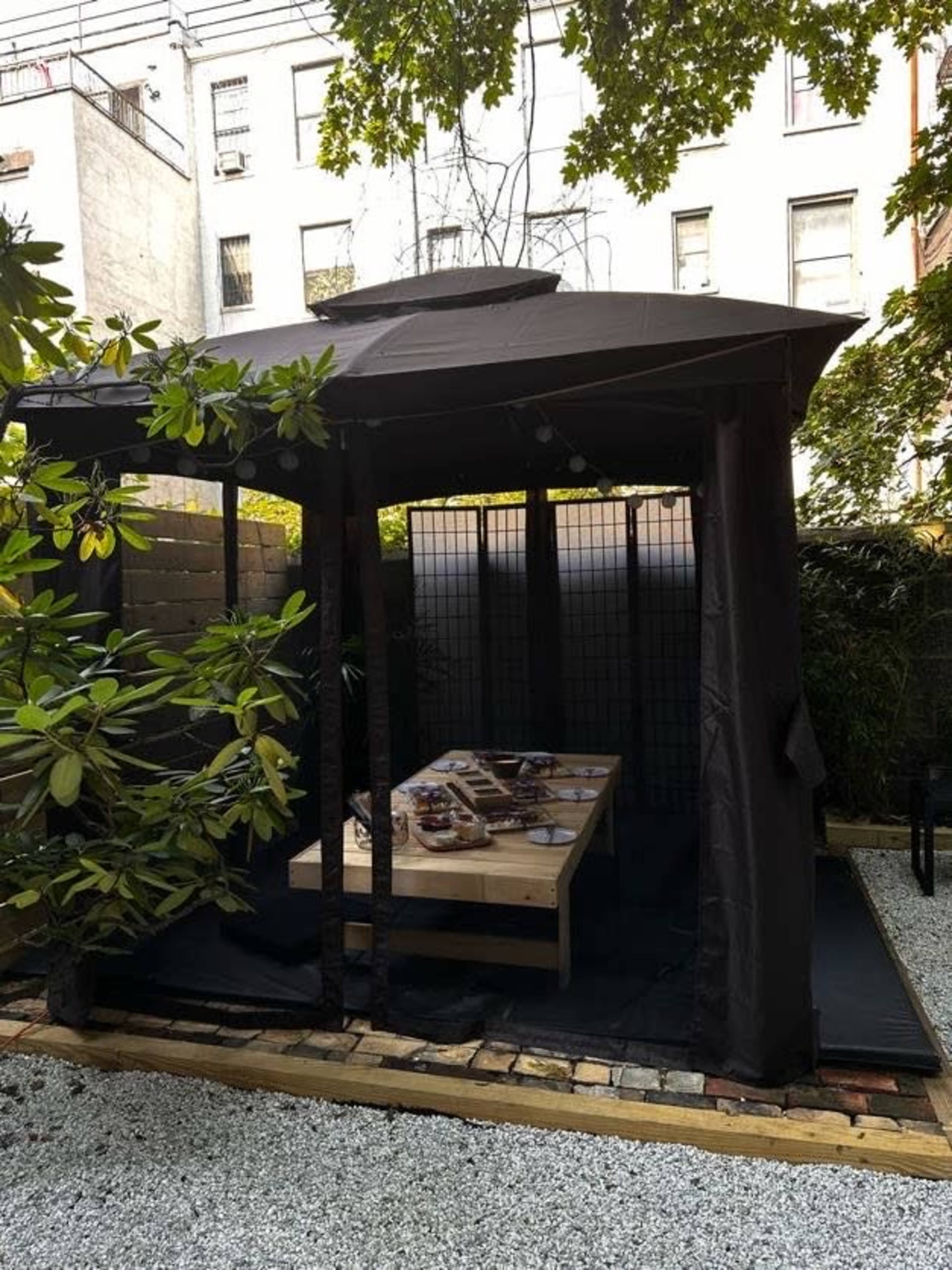 A dark canopy structure with a wooden table is situated in a gravel area surrounded by greenery.