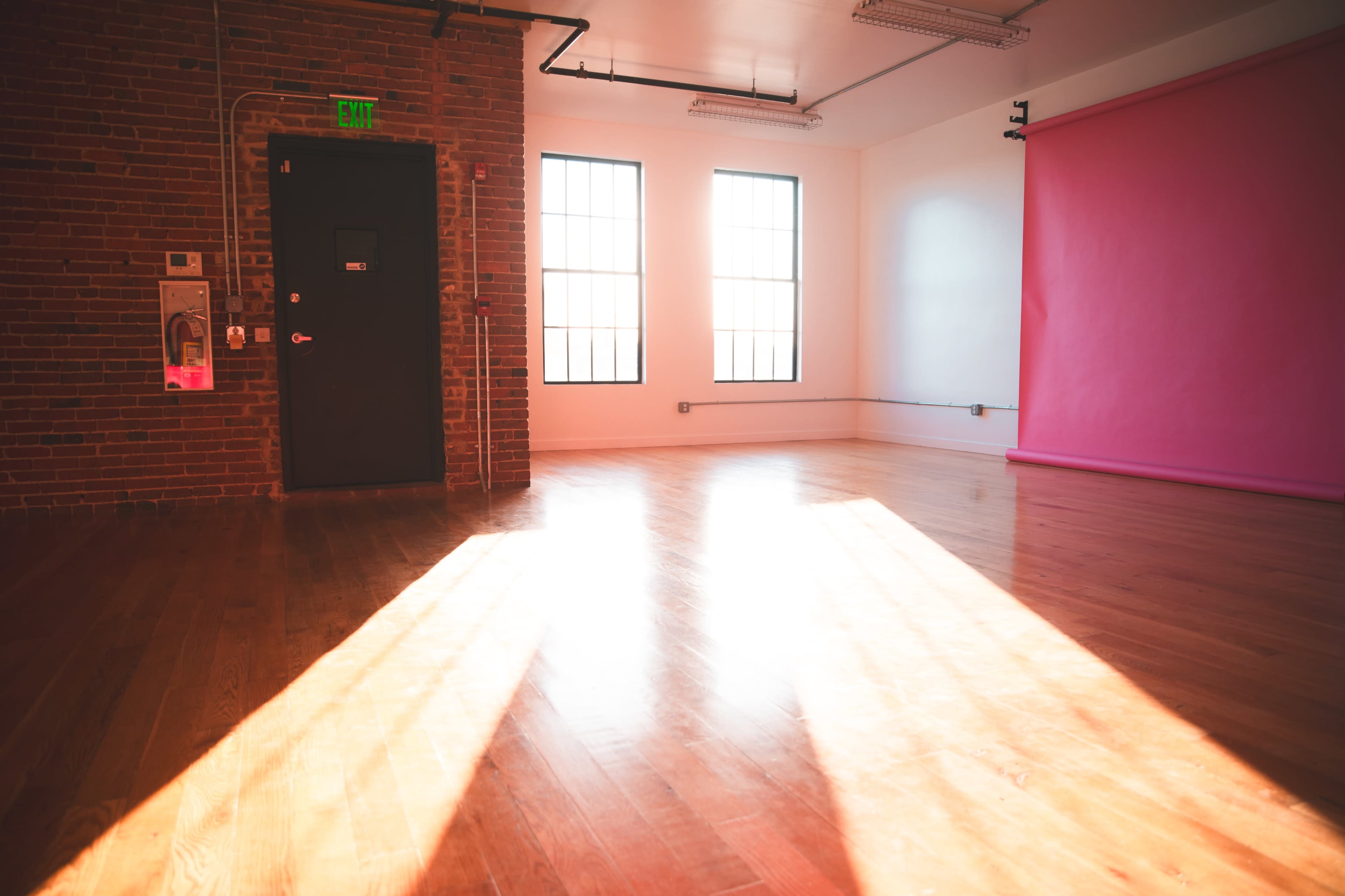 The image shows a spacious, well-lit room with wooden flooring, large windows, and a pink backdrop against one wall.
