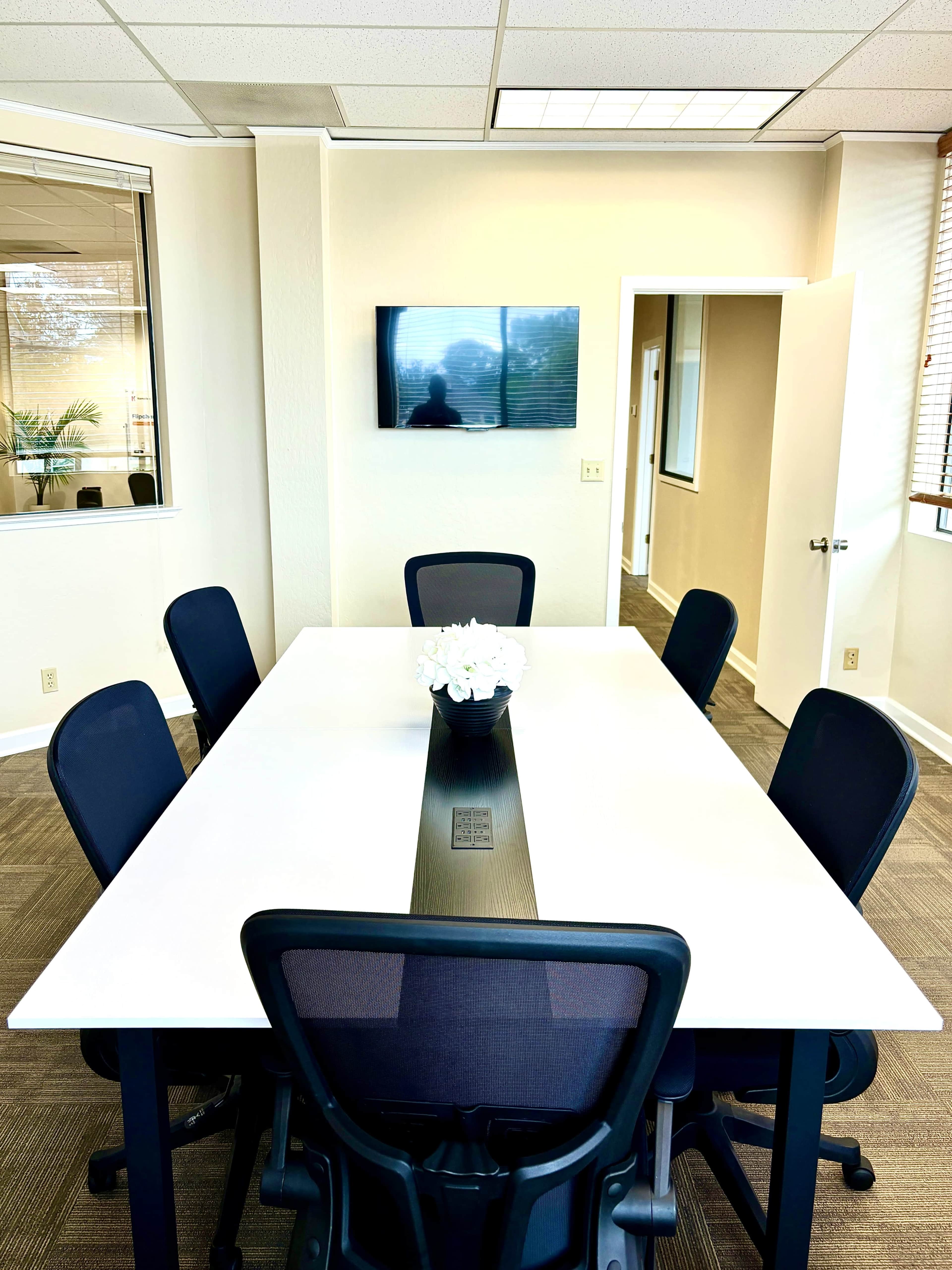 A modern conference room features a rectangular white table surrounded by black chairs, with a wall-mounted TV and a vase of flowers in the center.