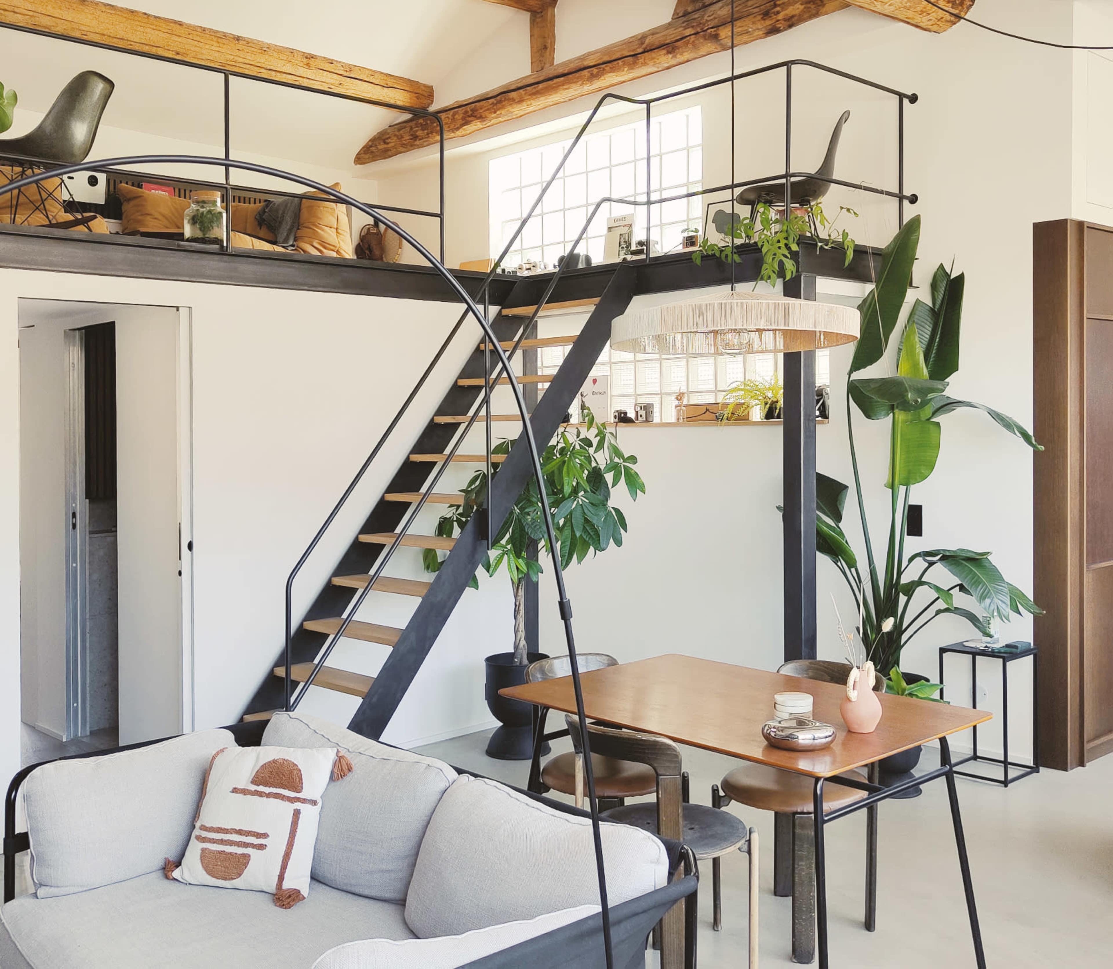 The image shows a modern living space with a staircase leading to a loft, a dining table, and decorative plants placed around the room.