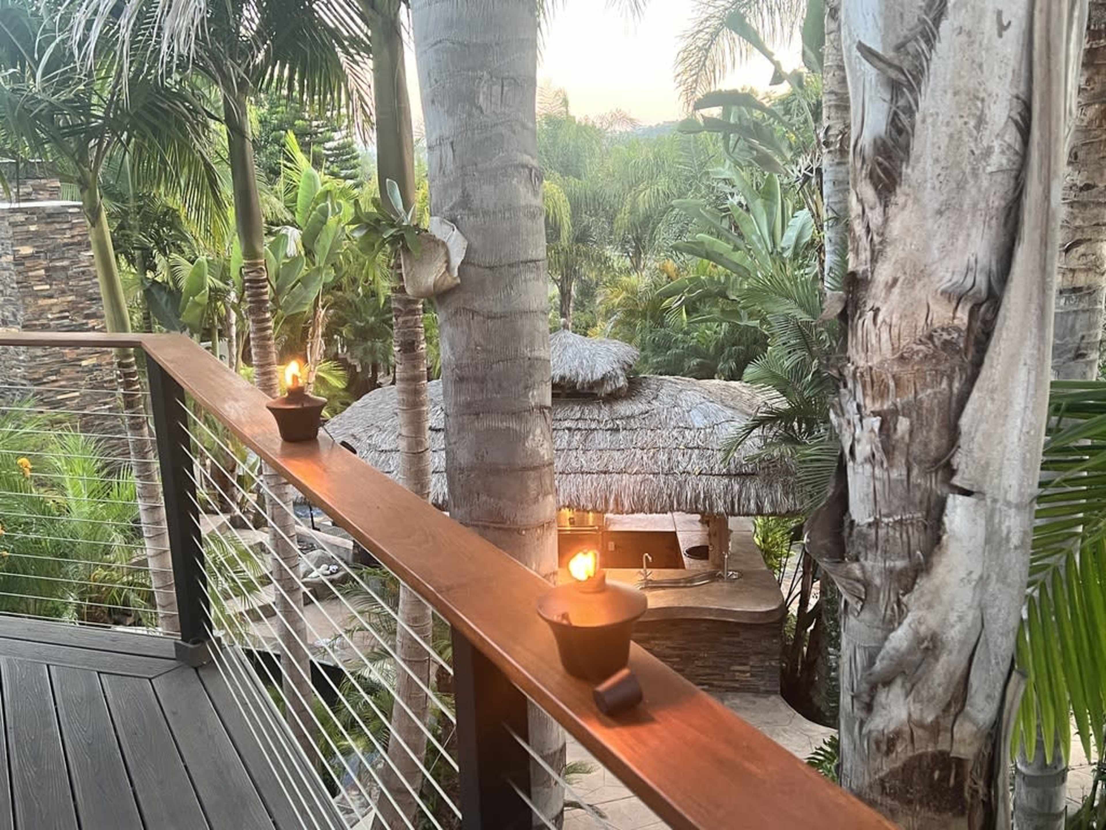 A balcony view with lit torches overlooking a tropical landscape and a thatched-roof structure among palm trees.