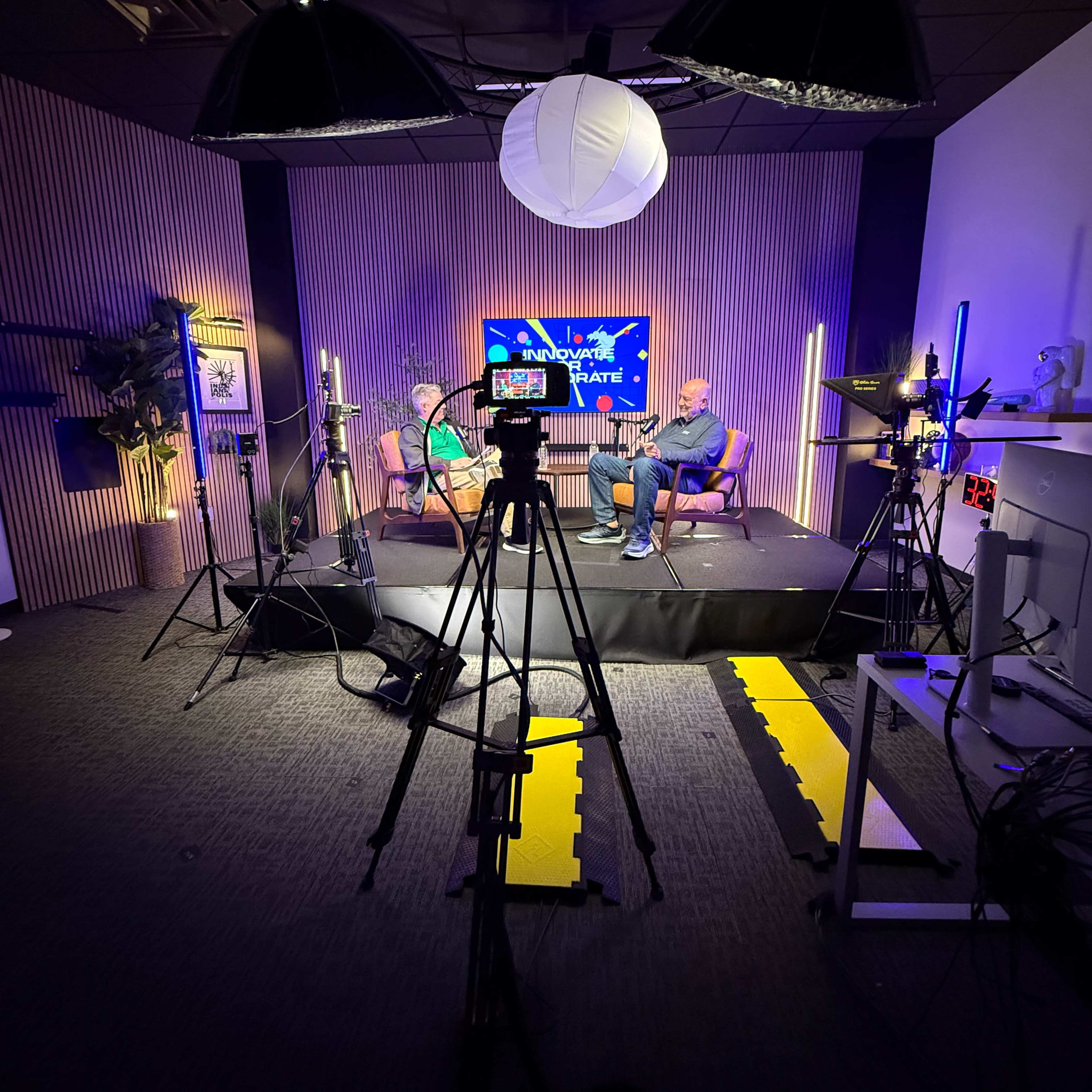 The image shows a studio set up for a video recording, featuring two chairs, multiple microphones, and lighting equipment, with a display screen in the background.