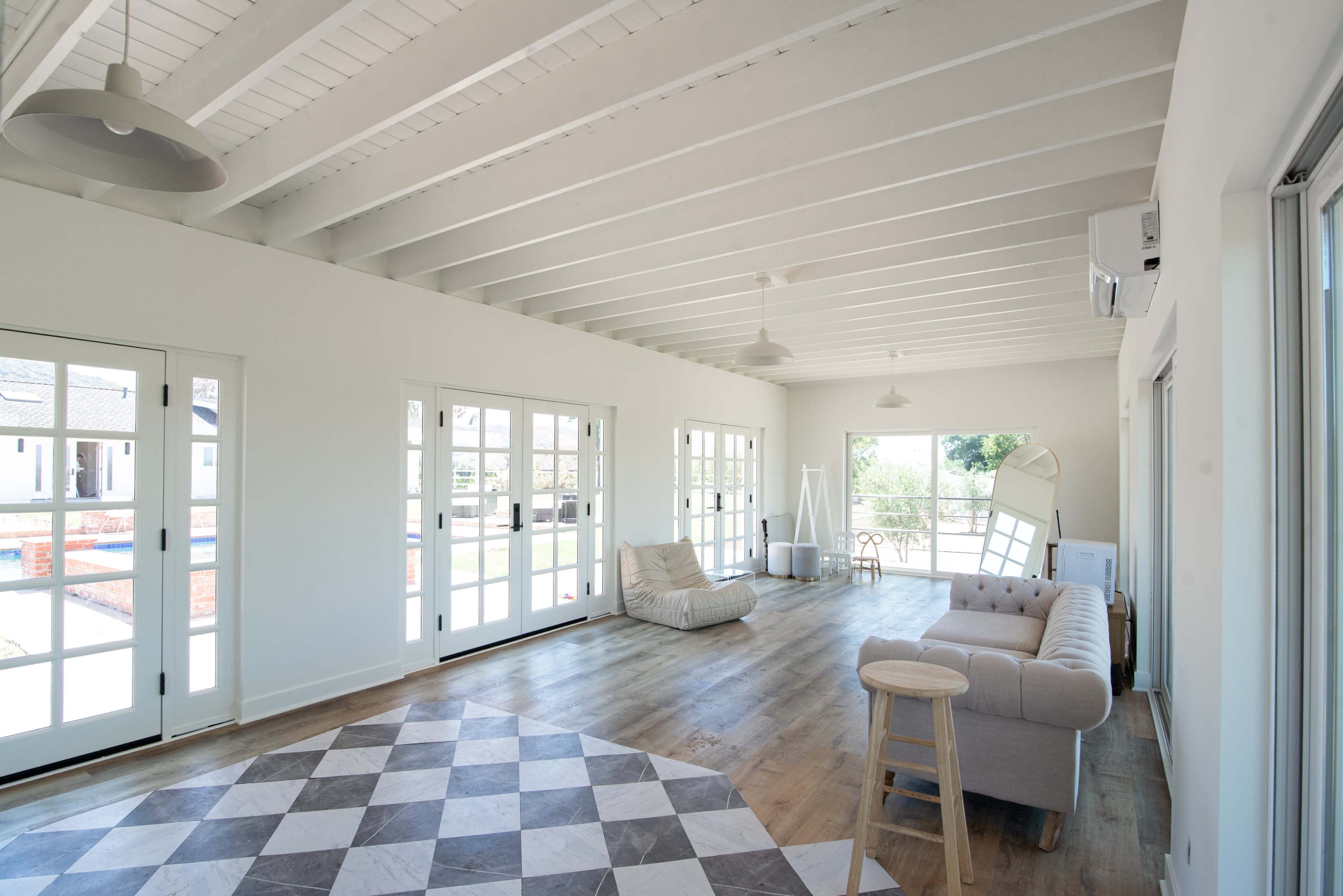 The image shows a spacious, modern interior with high white ceilings, large windows, and a light-colored sofa arranged on a patterned floor.