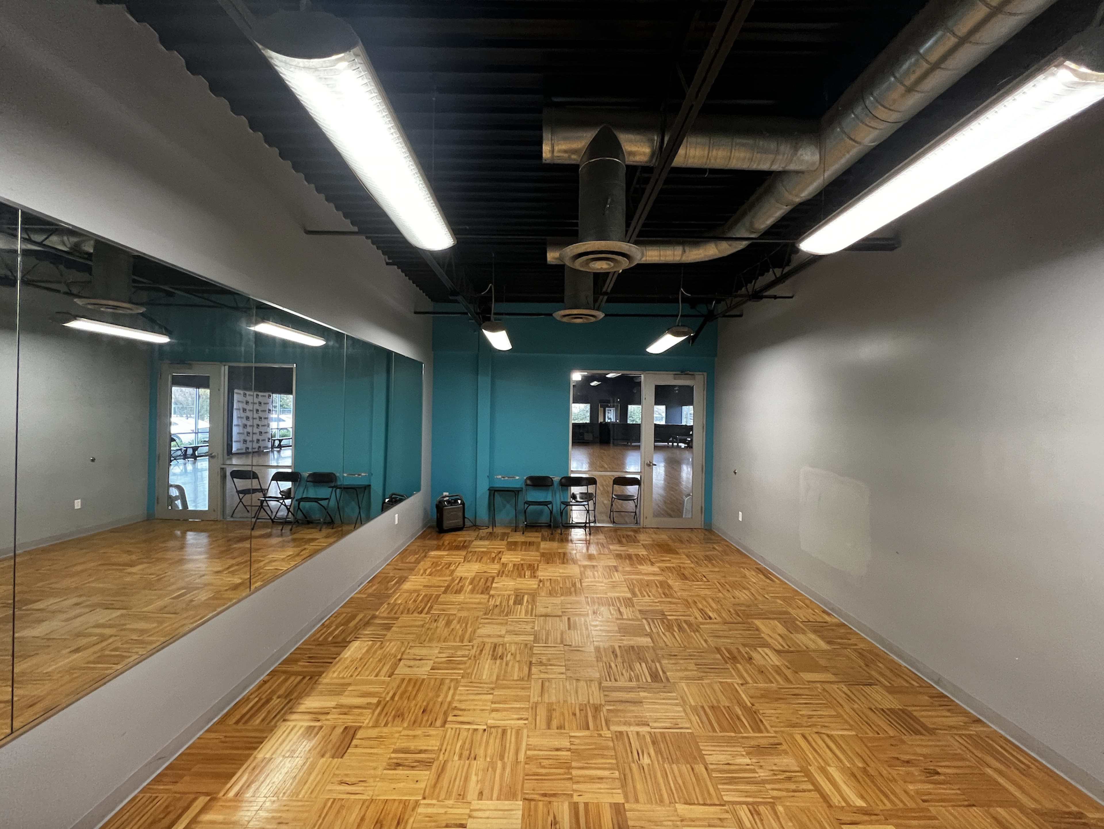 The image shows a spacious room with a wooden floor, two mirrors on one wall, and several chairs arranged near a blue accent wall.