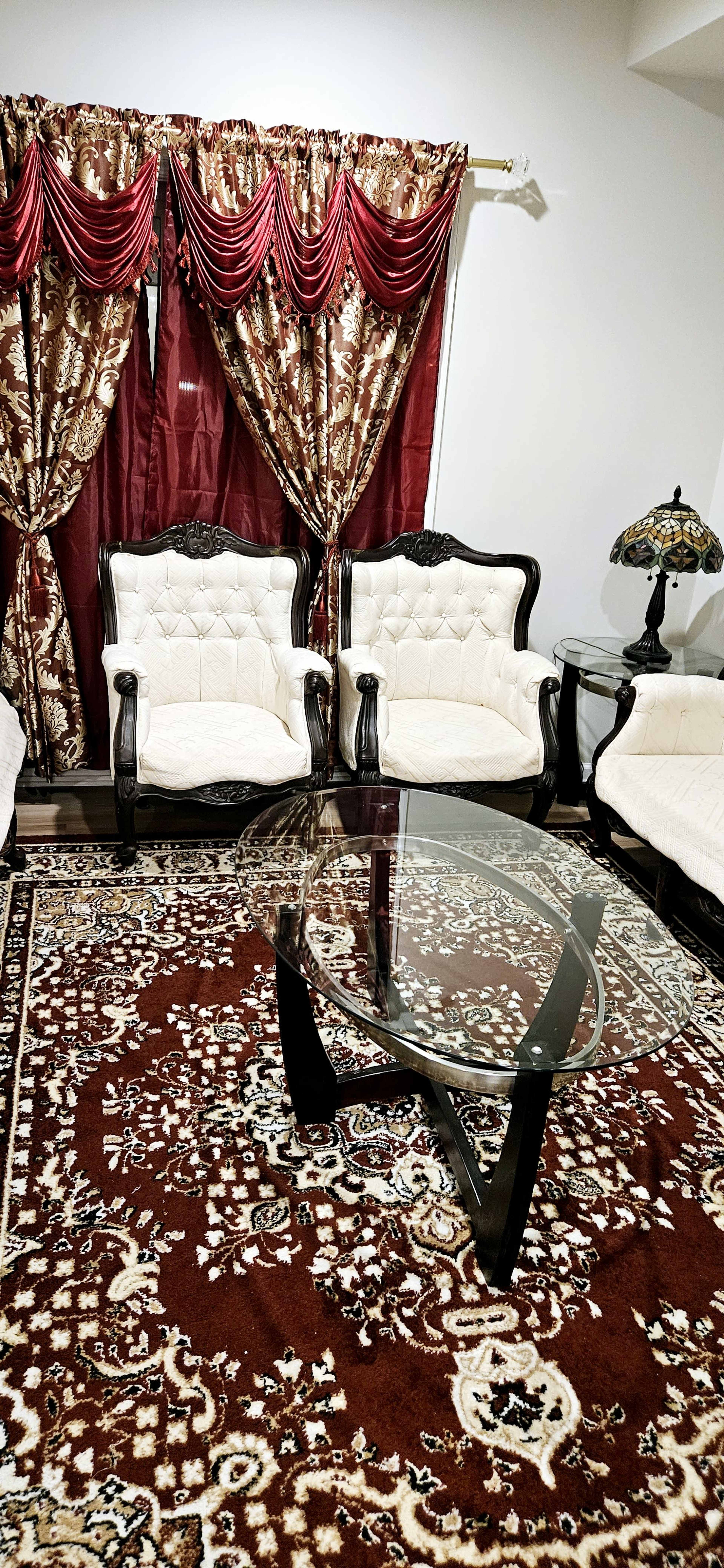 The image shows a living room with ornate upholstered chairs, a glass coffee table, and a decorative rug beneath.