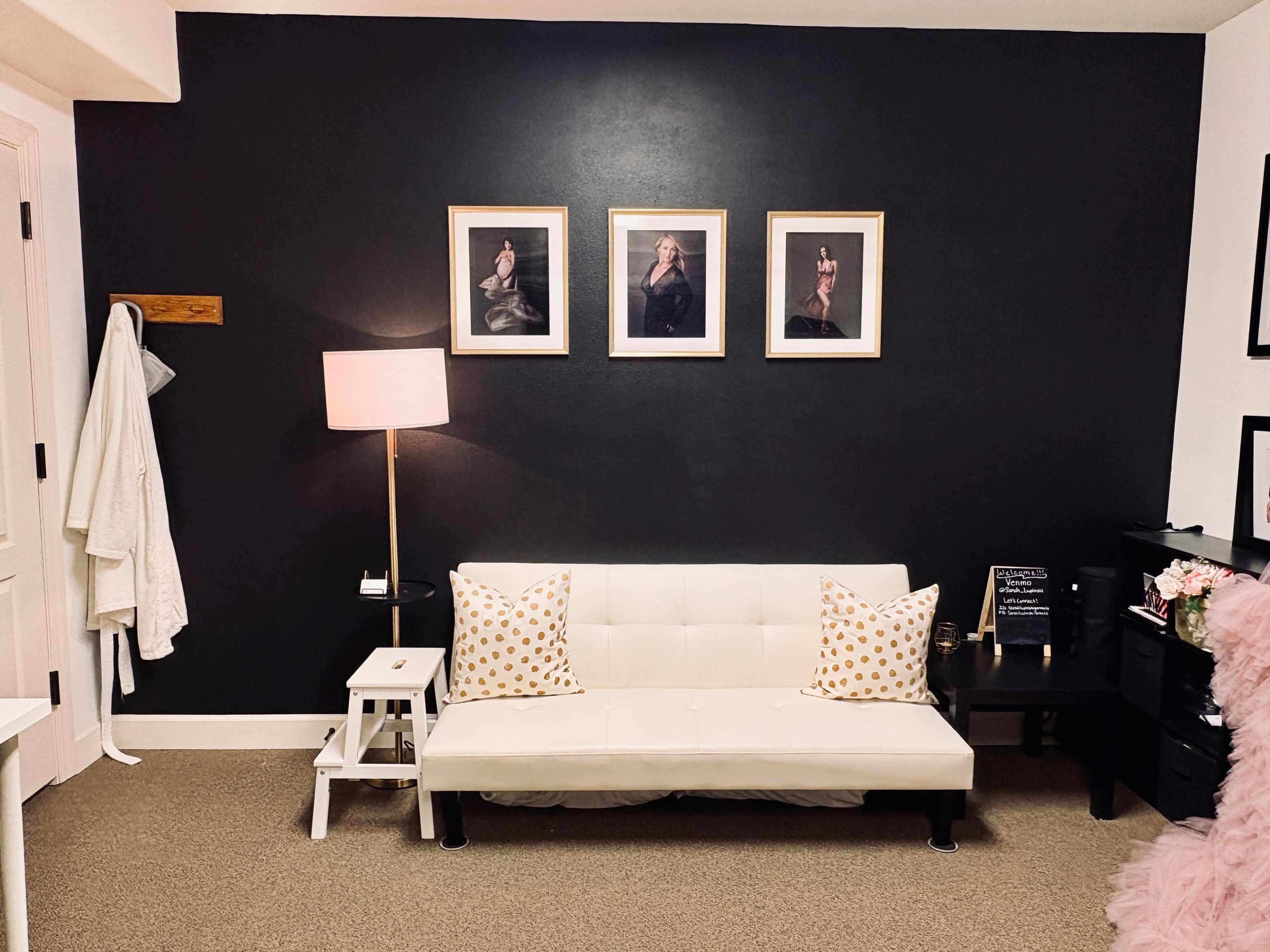 The scene features a small living area with a white futon adorned with patterned pillows, a standing lamp, framed photographs on the dark wall, and various decorative items nearby.