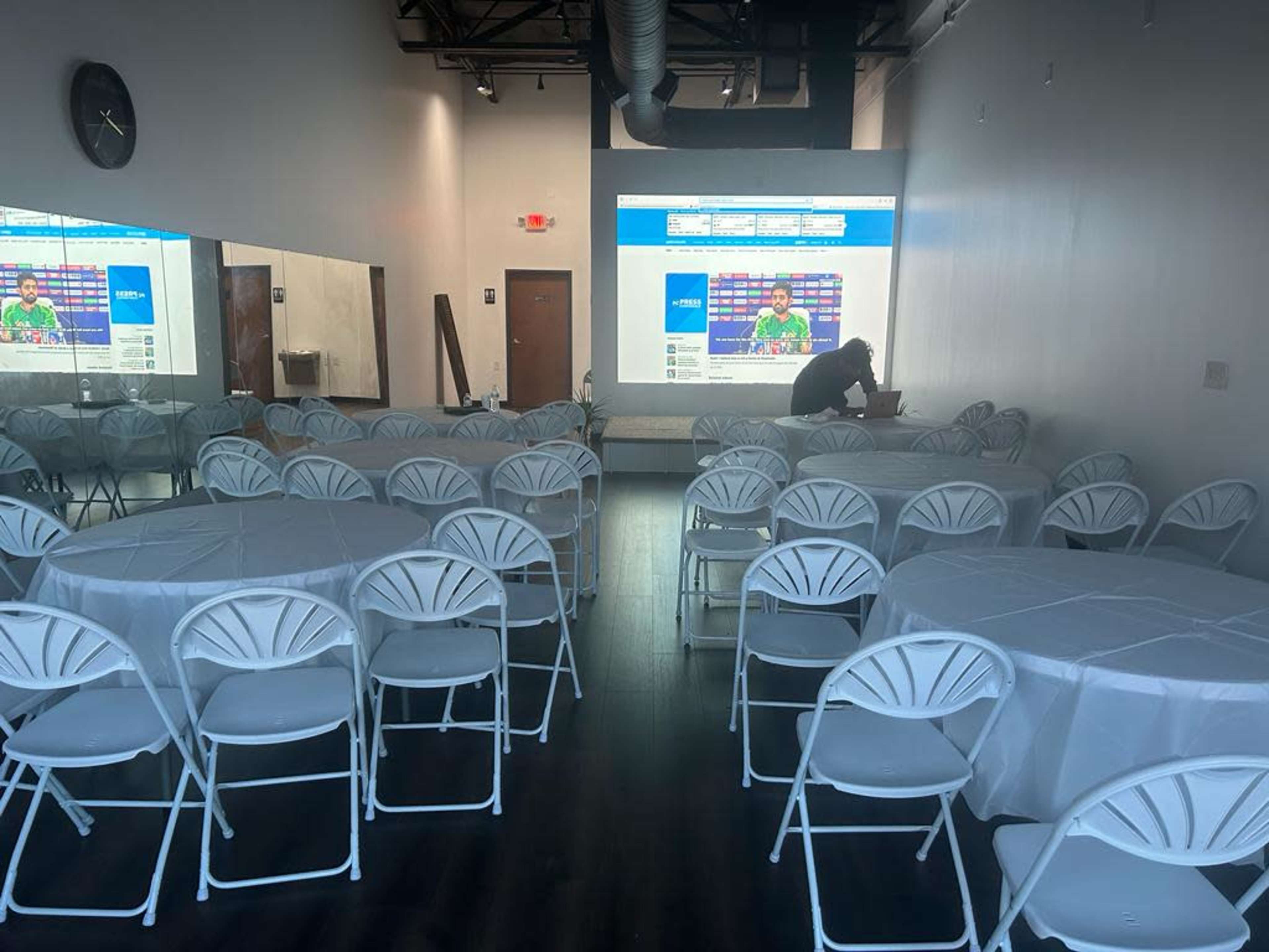 A spacious room set up with several round tables and white chairs, with a large screen displaying information at the front.