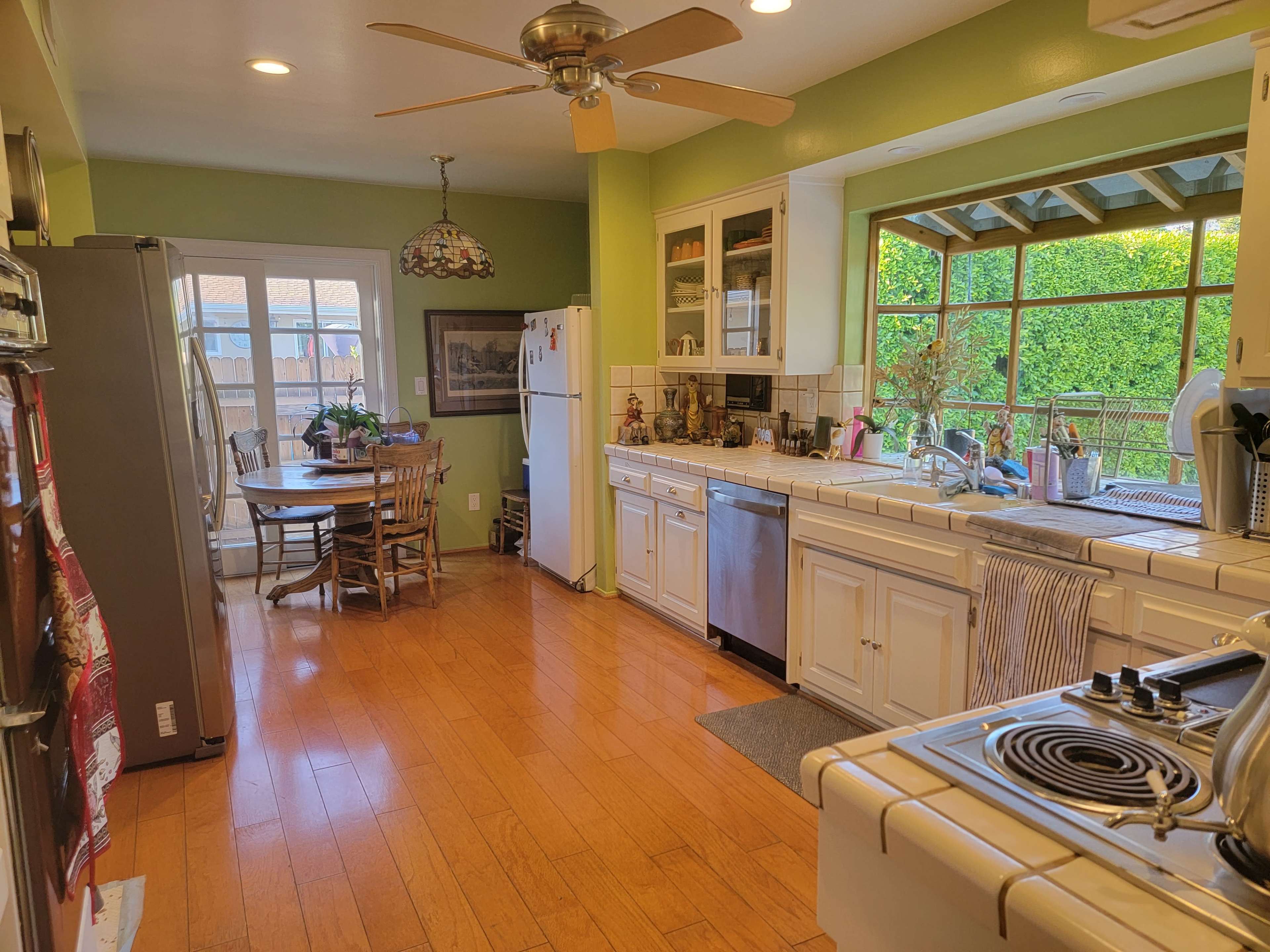 A bright kitchen features white cabinetry, a central island with a stove, and an eating area with a round table and chairs, all illuminated by natural light from a large window.