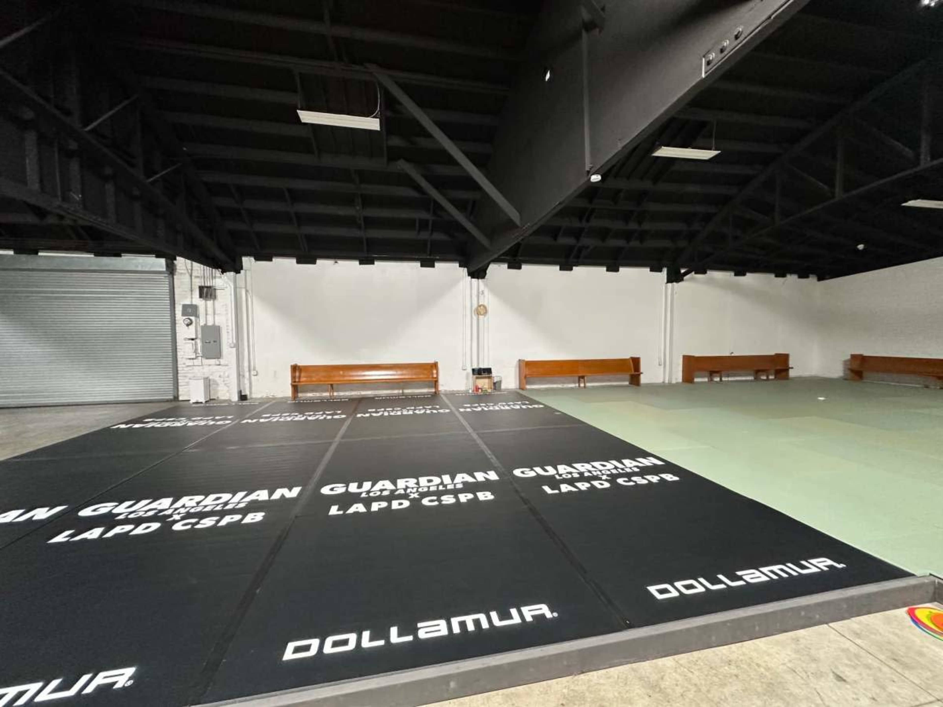 The image shows a spacious indoor area with black and green mats, benches along the walls, and a high ceiling.