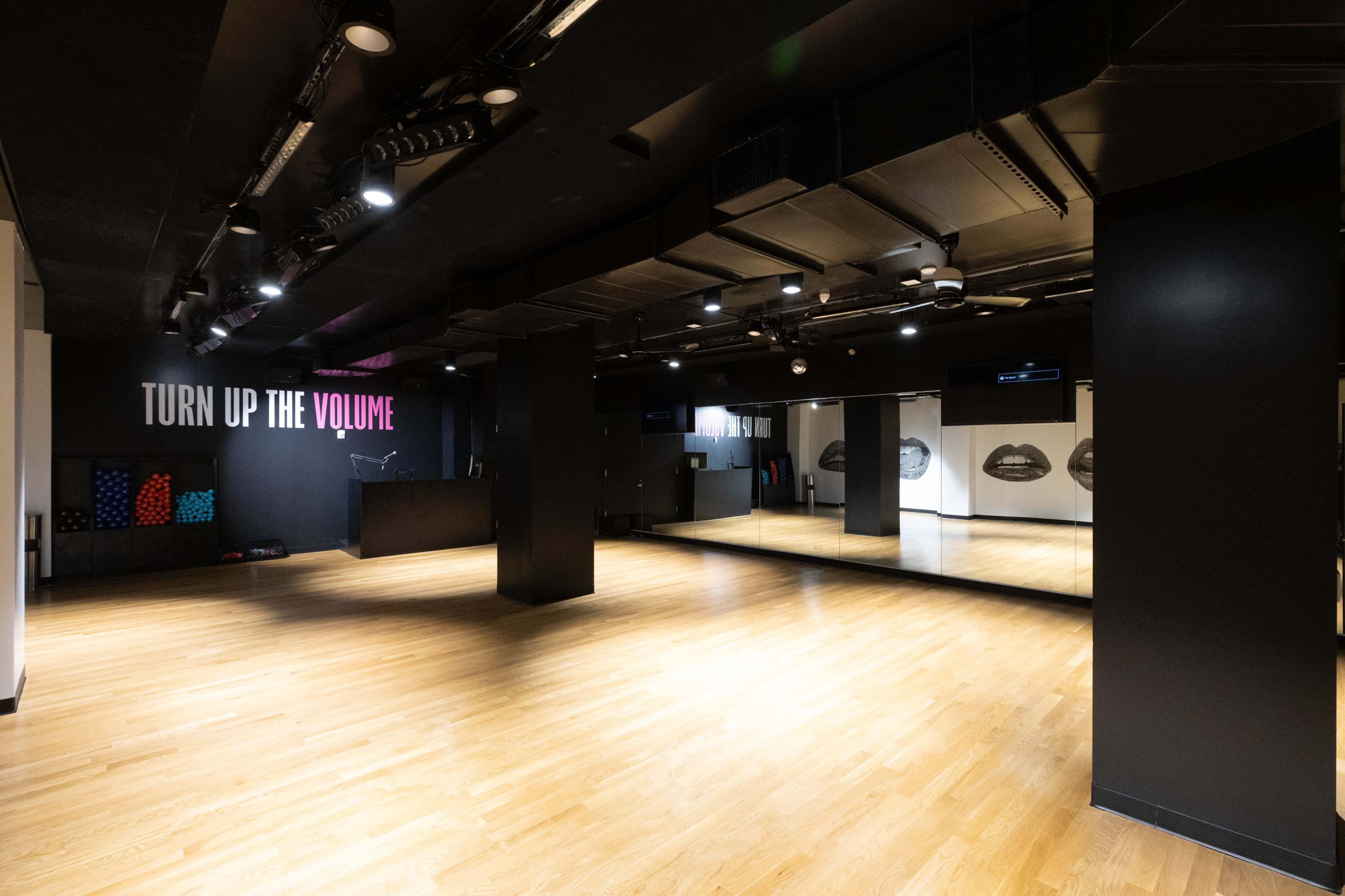 A spacious, modern studio with wooden flooring, black walls, and large mirrors, featuring a "TURN UP THE VOLUME" sign.