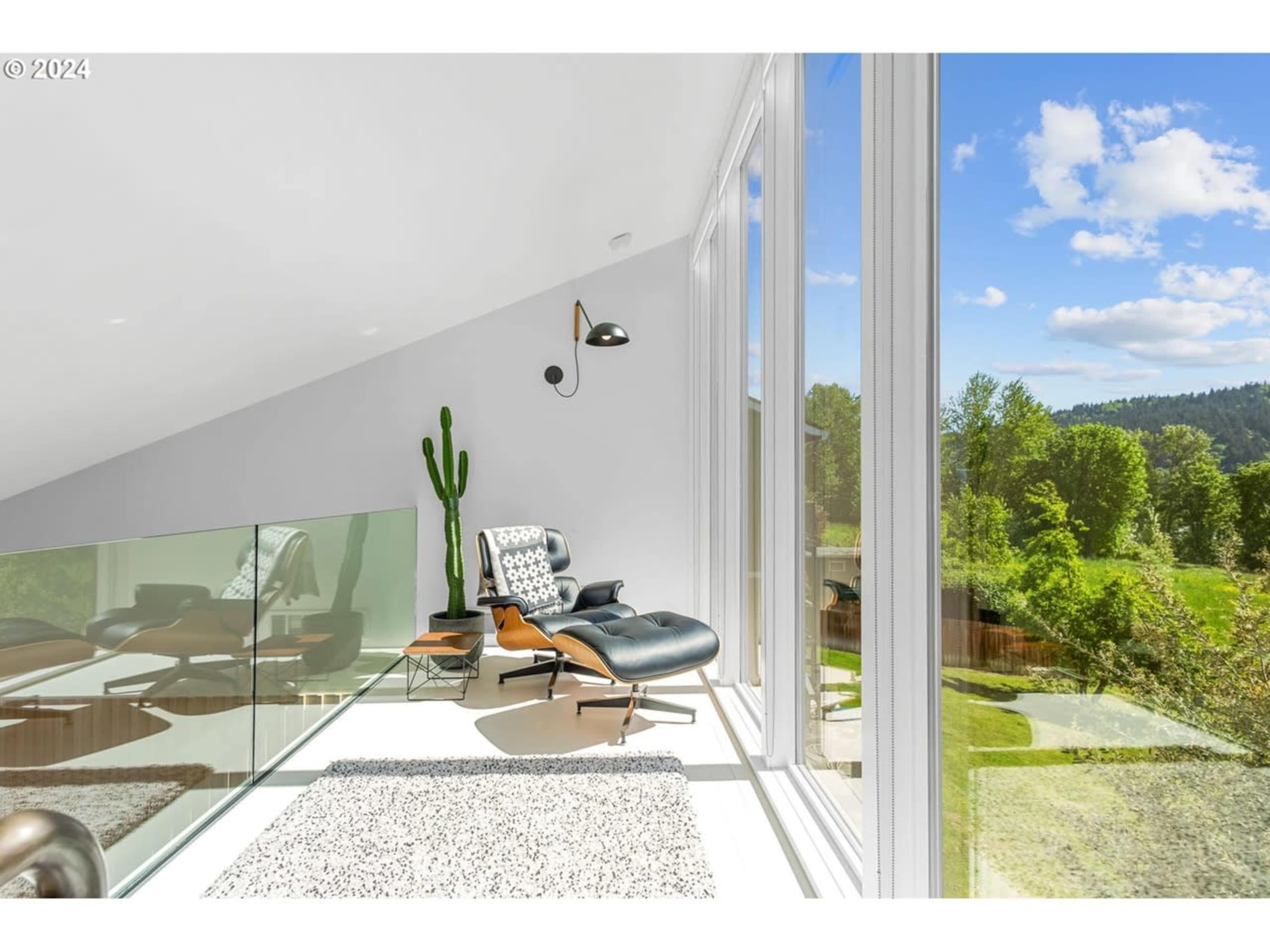 A modern lounge area features an Eames chair and a cactus by large windows overlooking greenery and a sunny sky.
