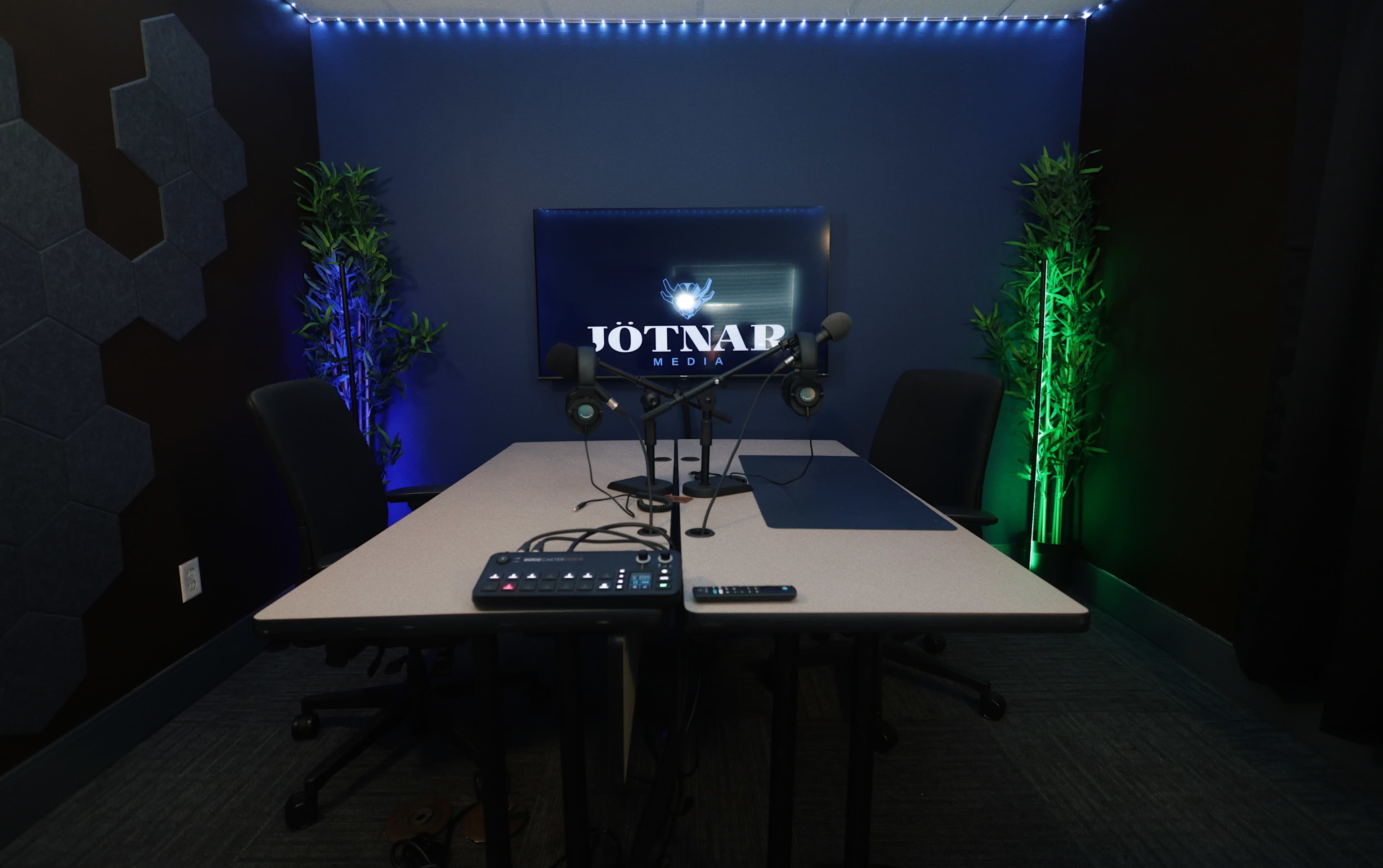 The image shows a recording studio with a table, microphones, and a large screen displaying the logo of Jotnar Media, surrounded by subtle lighting and greenery.
