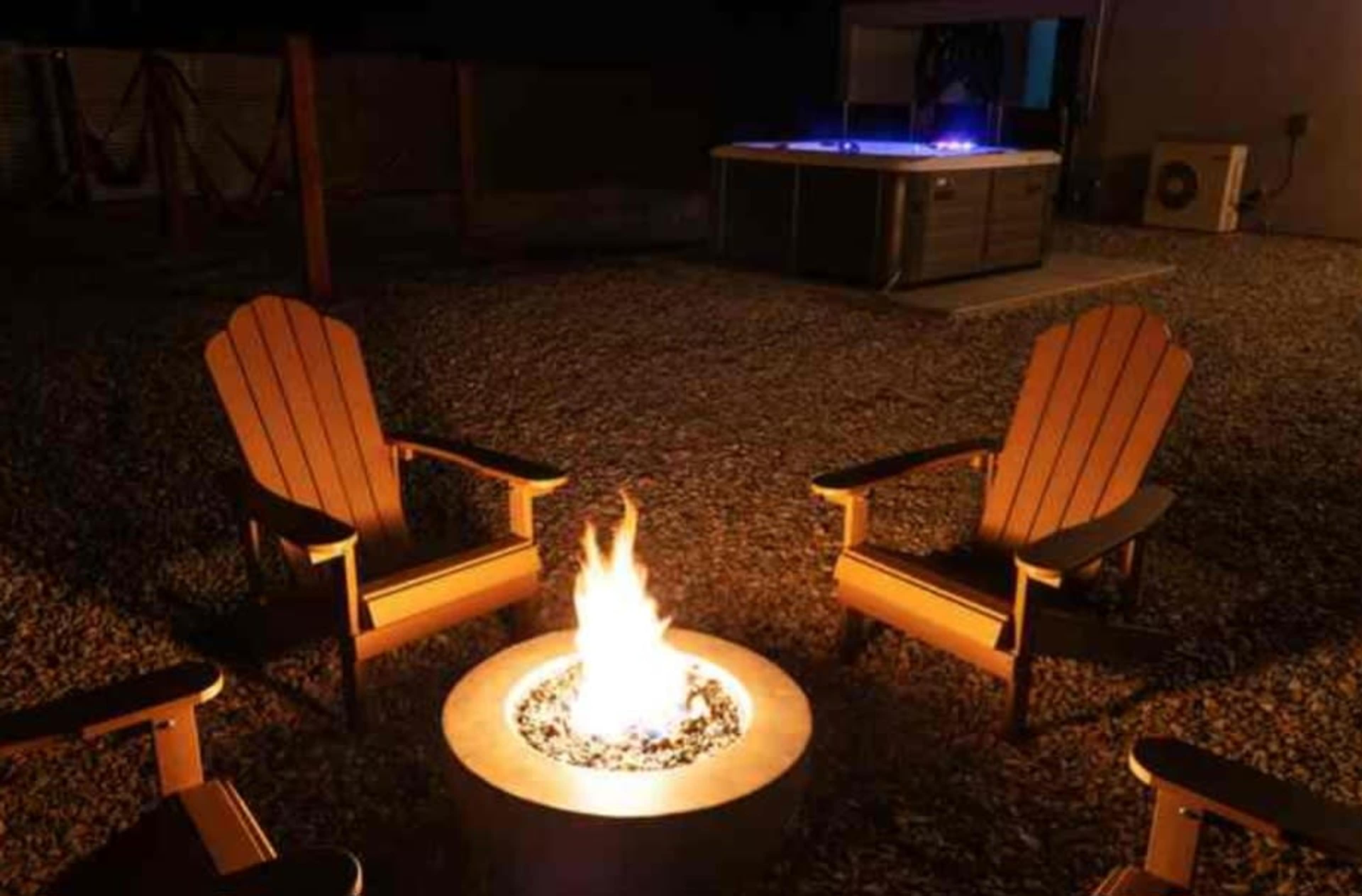 A fire pit surrounded by four wooden chairs is illuminated at night, with a hot tub visible in the background.