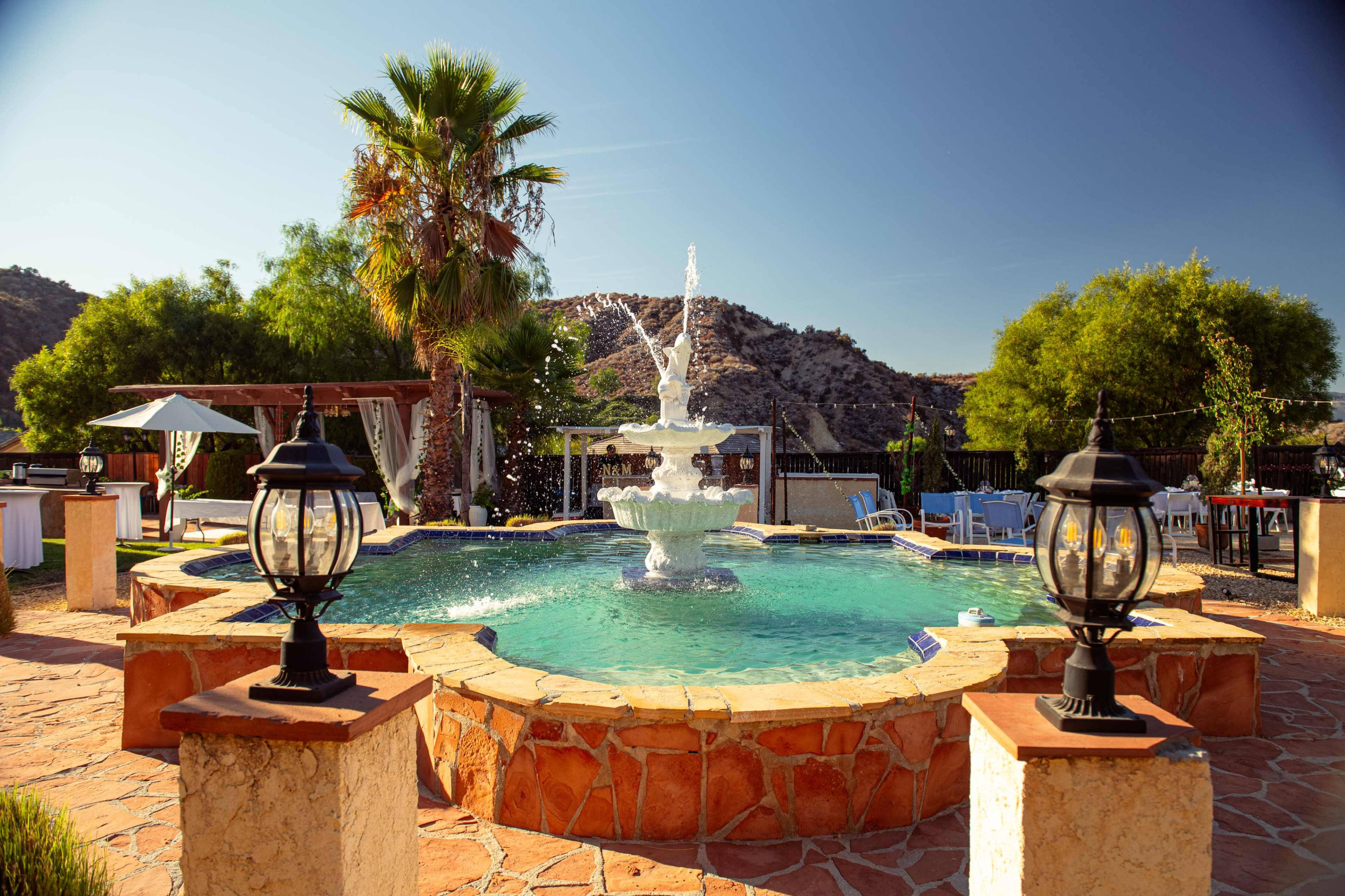 The image shows a circular fountain surrounded by a pool, with green trees and mountains in the background, and decorative lanterns positioned around the stone border.