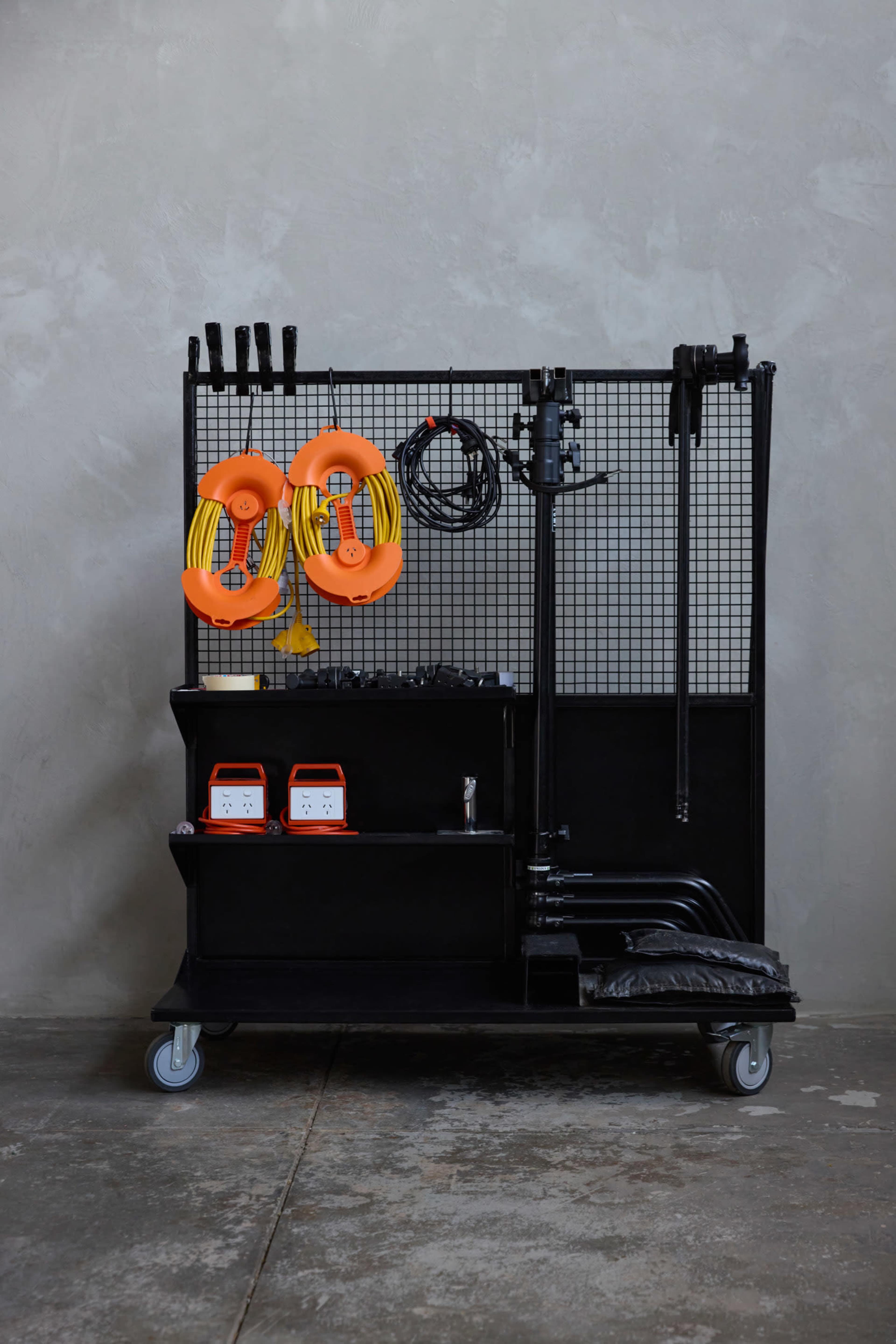 A black rolling cart with various tools and equipment organized on a metal grid, including two orange reels, wires, and battery-powered devices.