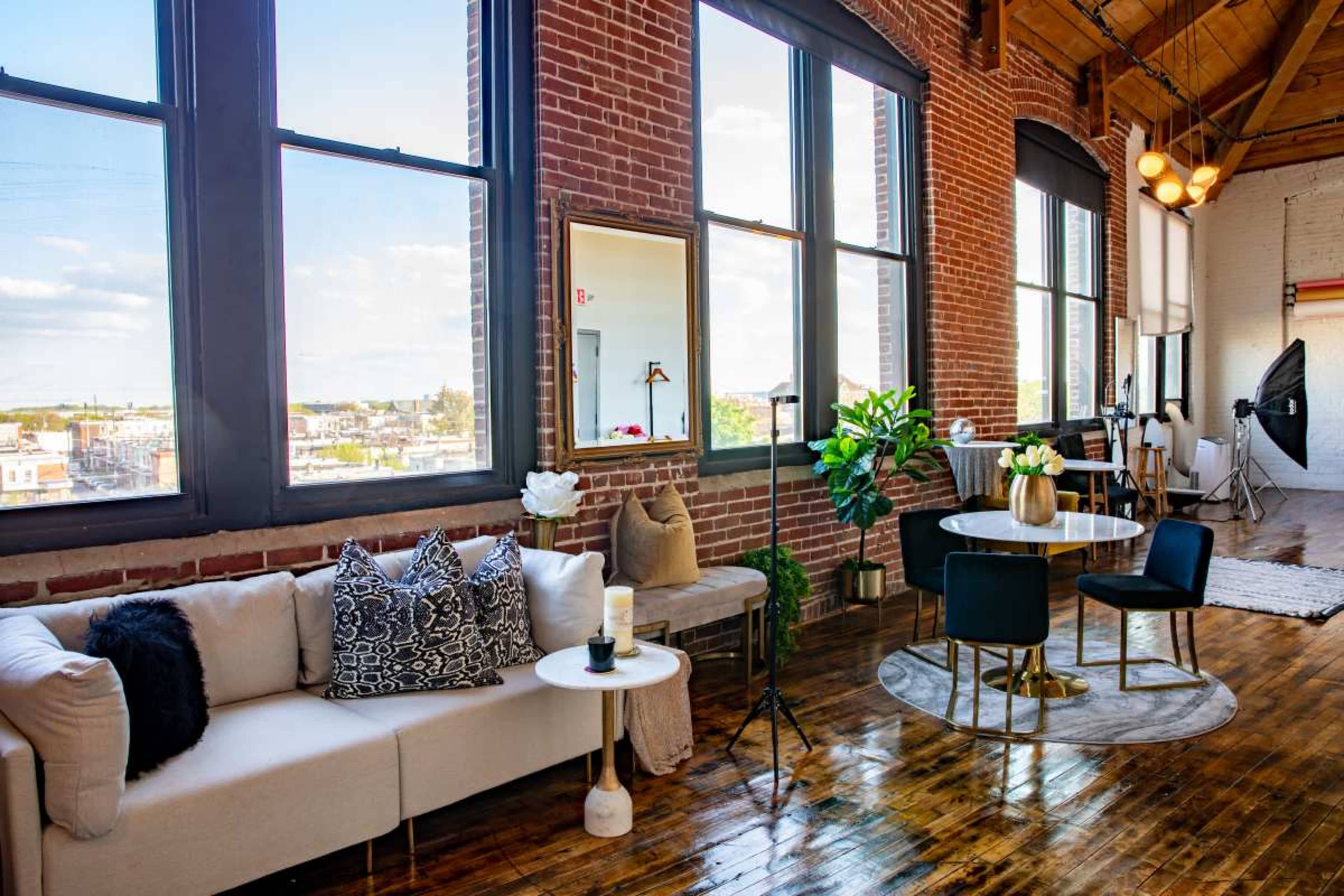 The image shows a stylish loft interior with large windows, exposed brick walls, a light-colored sofa, a round dining table, and wooden flooring.