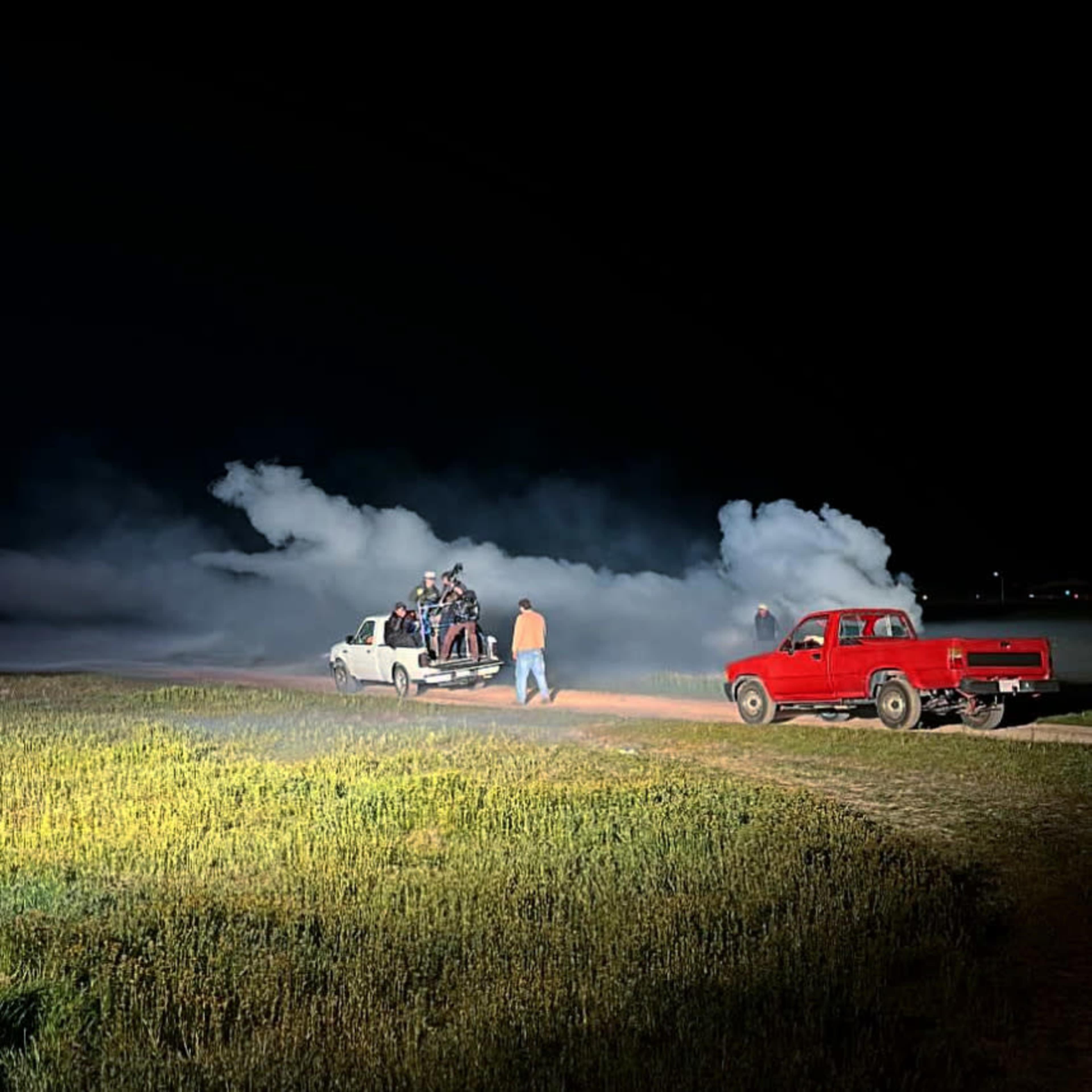 A group of people is gathered around a pickup truck on a dirt road at night, with smoke rising in the background.