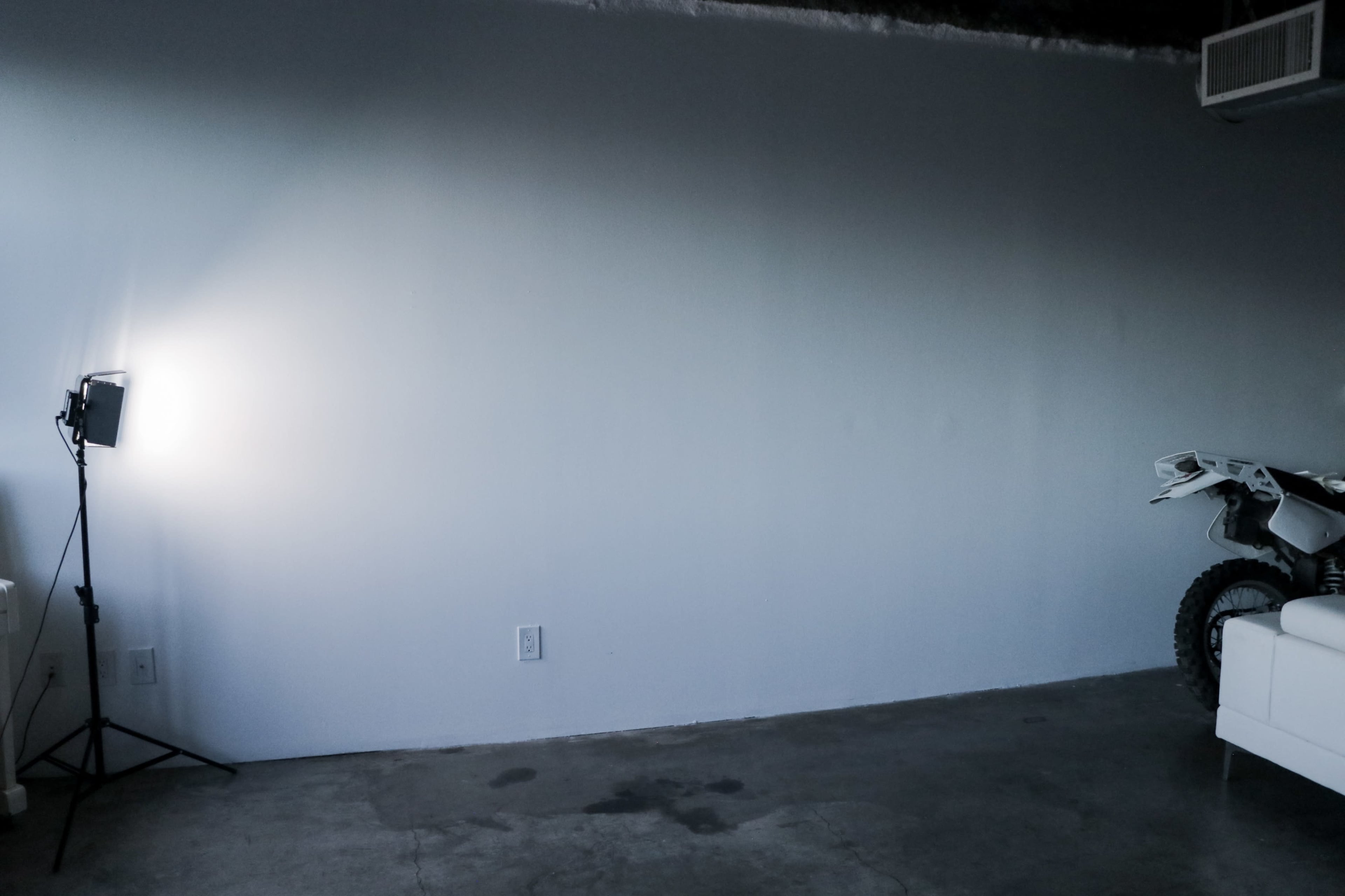 A sparsely furnished room with a white wall, a light source to the left, and a dirt bike partially visible on the right.