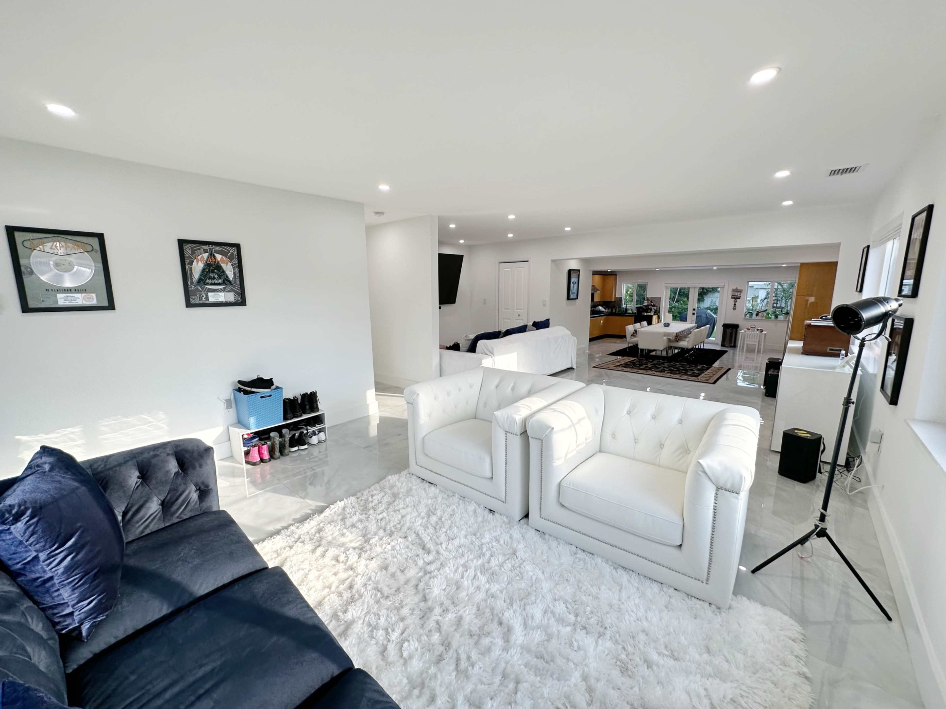 A spacious living room features a white rug, two white armchairs, a blue couch, and an open layout connecting to a kitchen area in the background.
