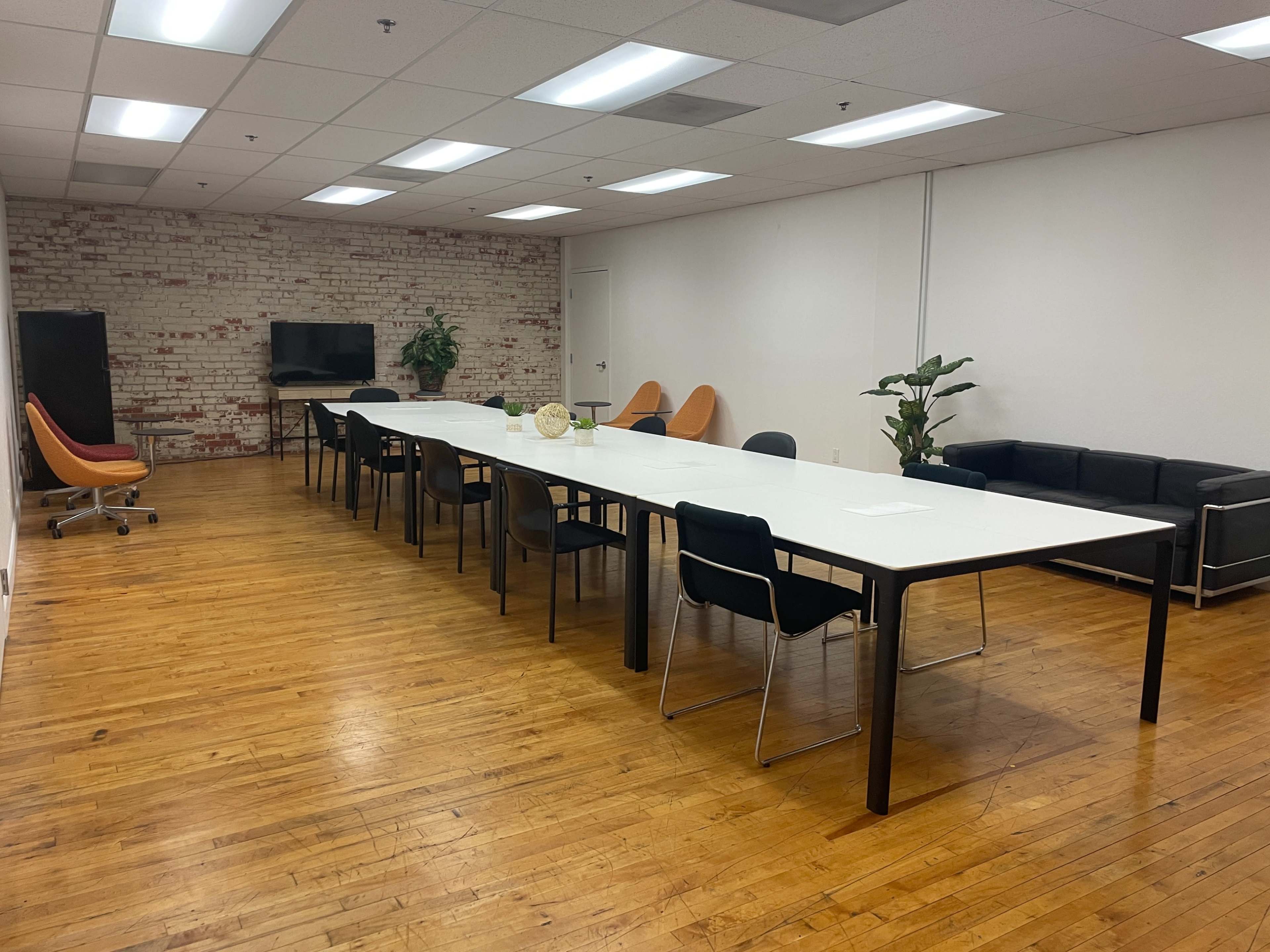 A spacious meeting room with a long table surrounded by chairs, a couch, a television, and decorative plants against a brick wall.