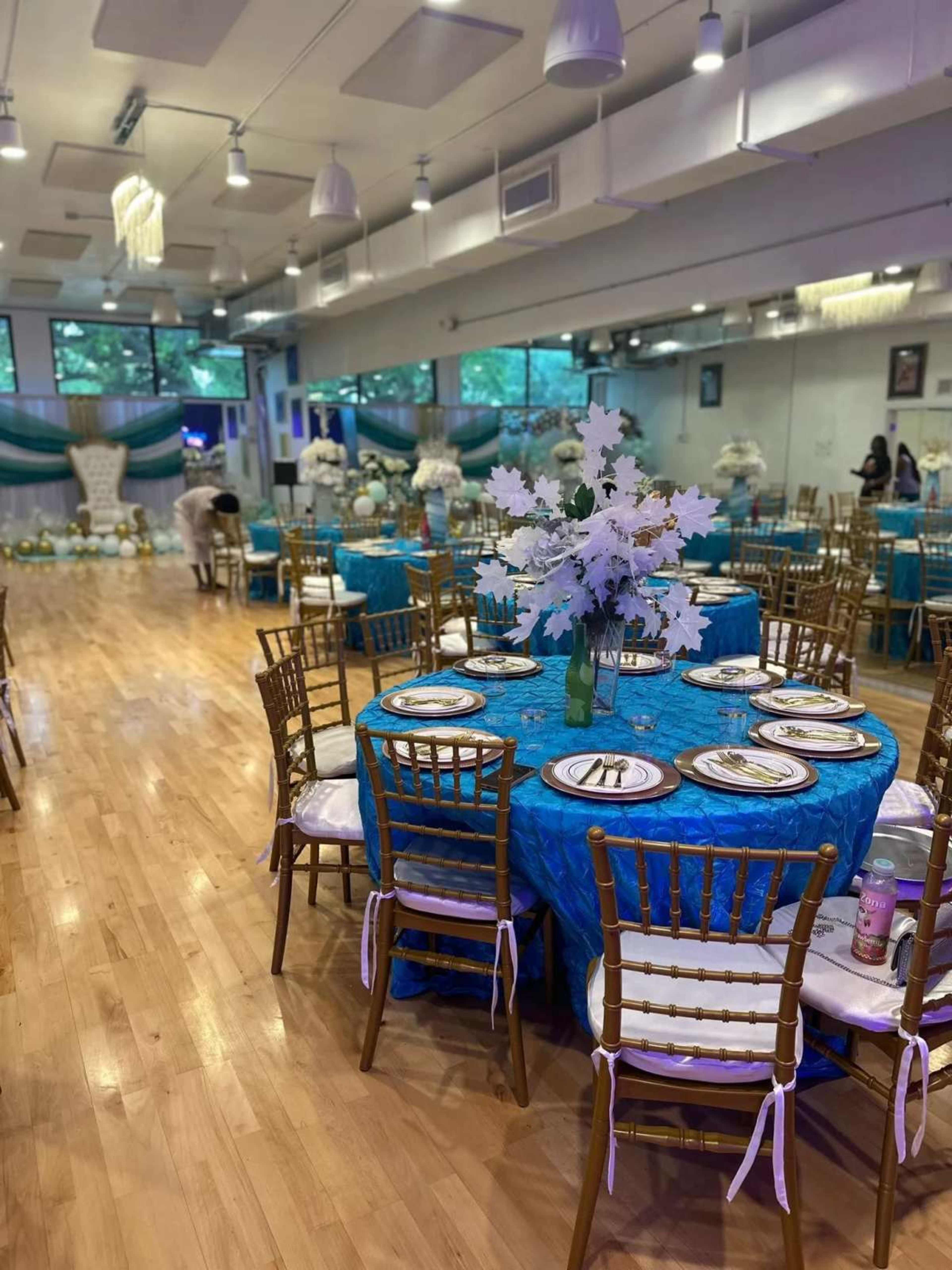The image shows a decorated event space featuring round tables with turquoise tablecloths, golden chairs, and floral centerpieces, set up for a formal gathering.