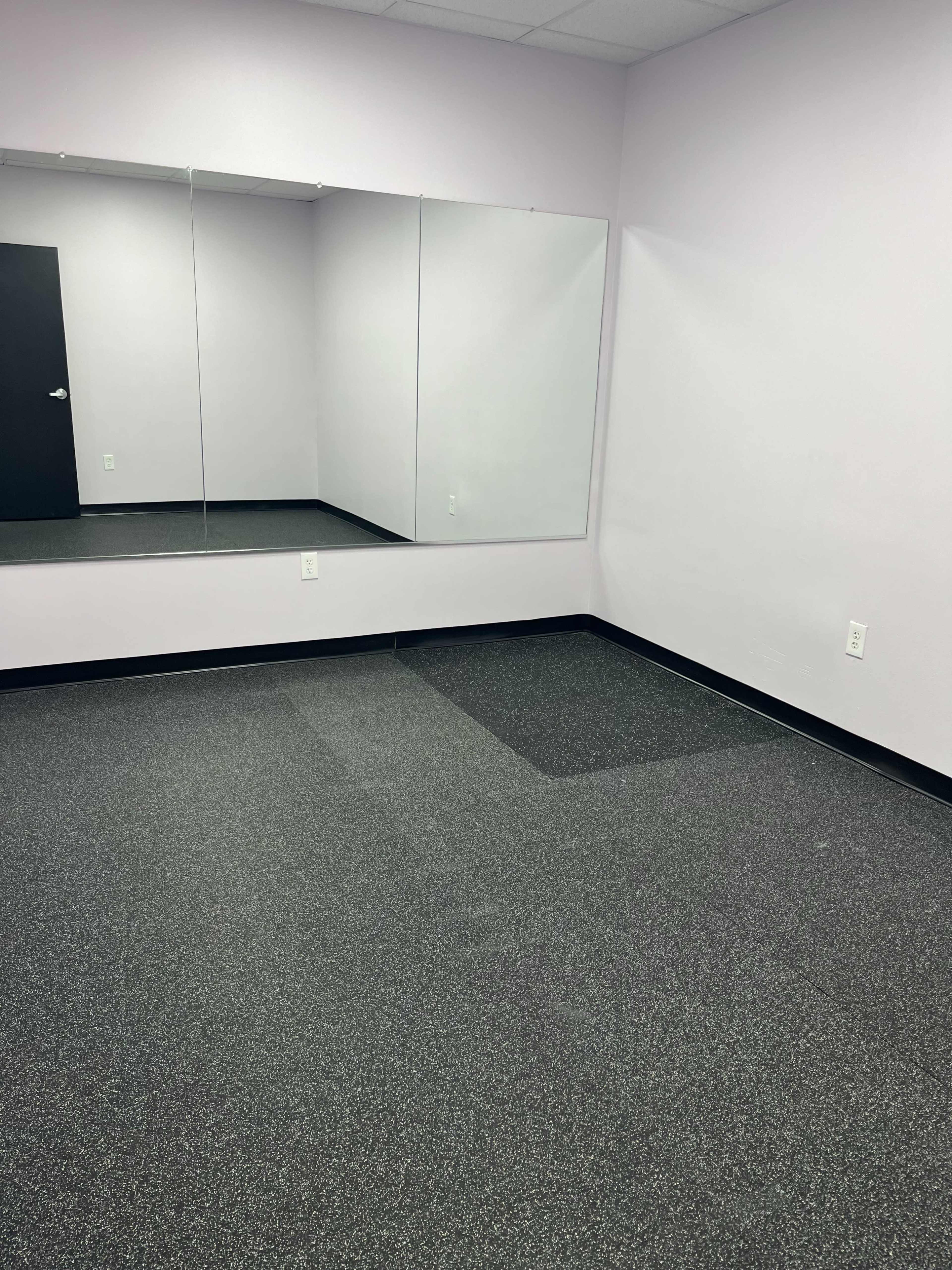 The image shows a sparsely furnished room with light purple walls, black flooring, and a wall-mounted mirror.