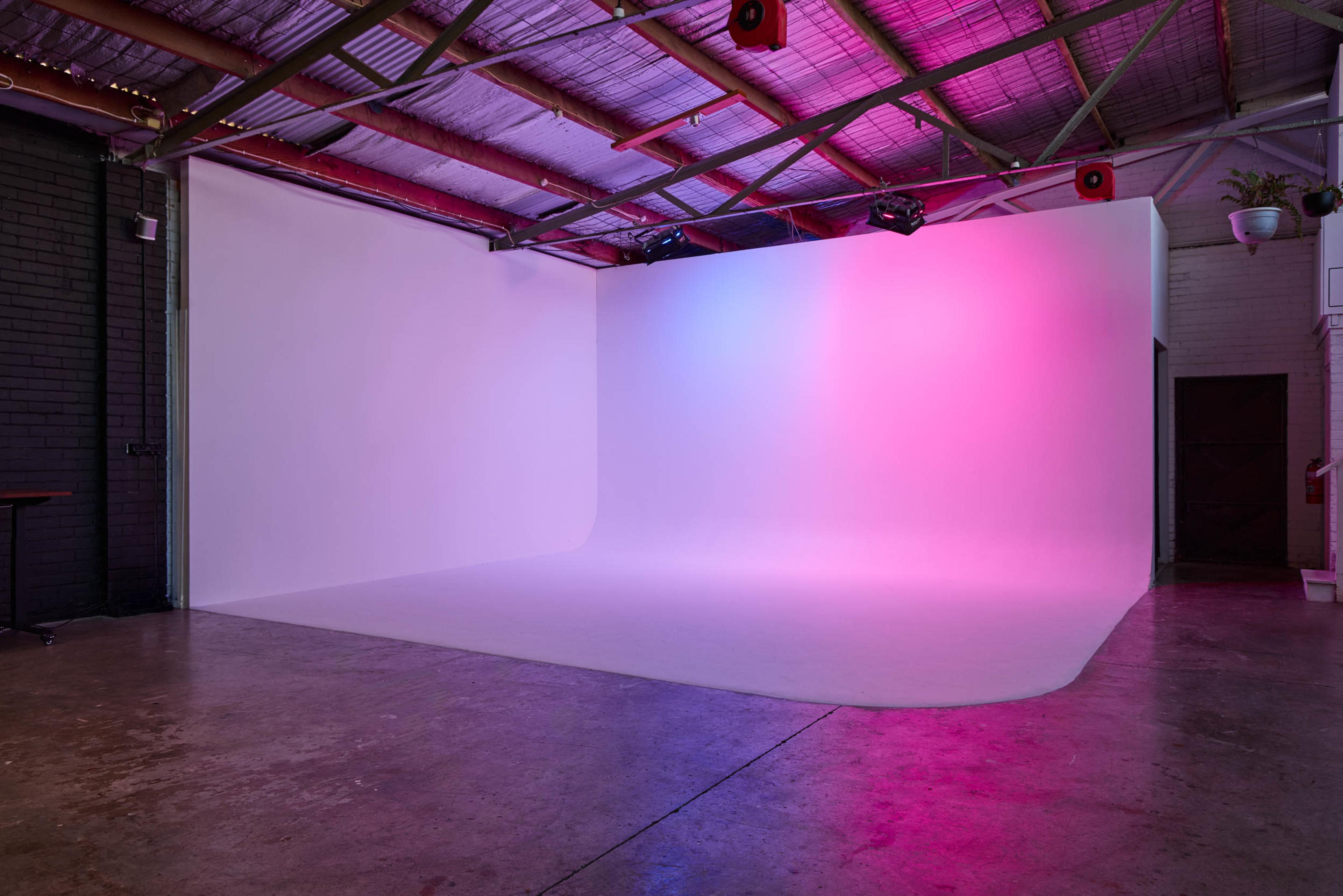 The image shows a photography studio with a curved white backdrop illuminated in pink and purple lighting.