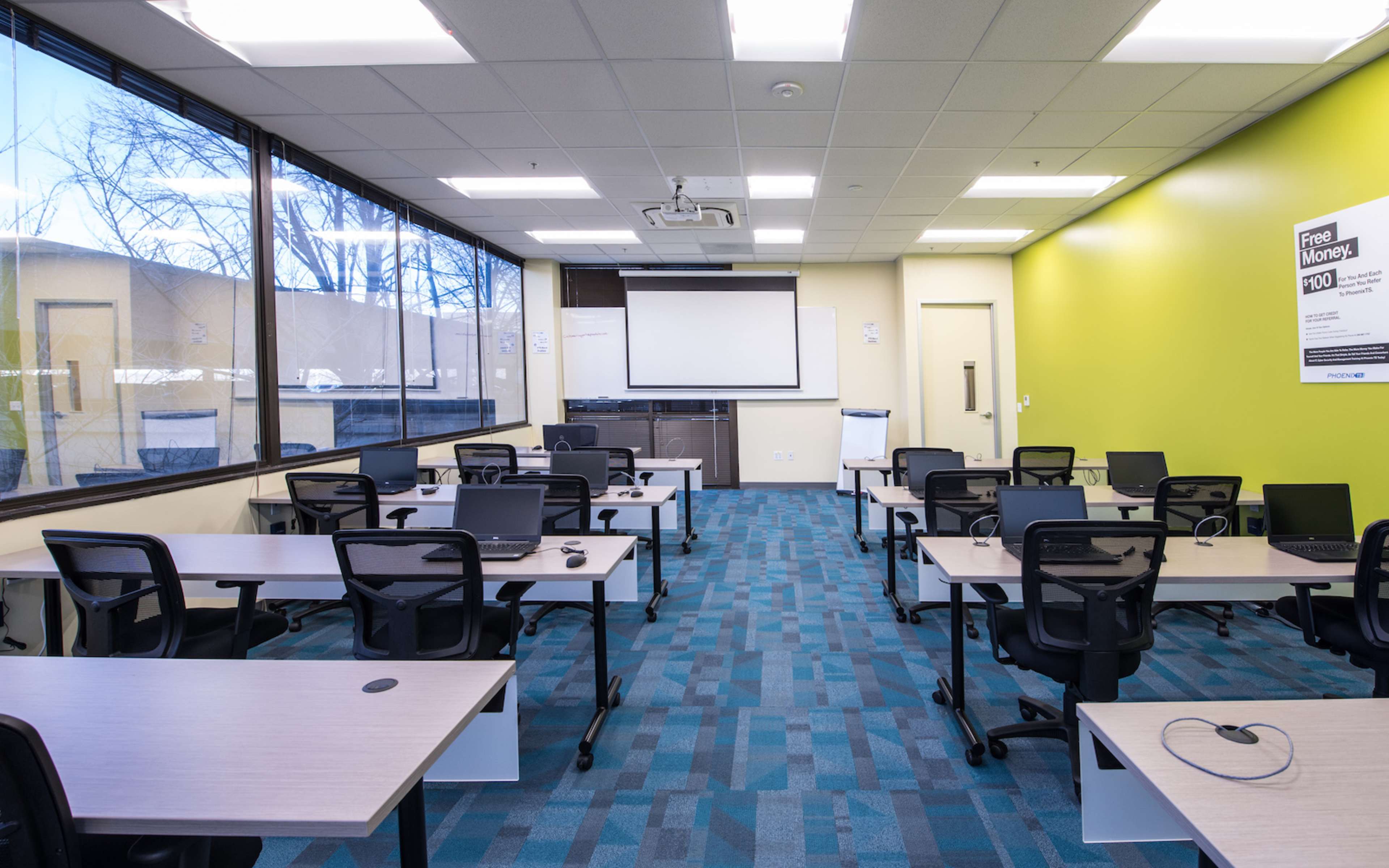 A modern classroom features several desks with computers, arranged for a training session, along with a projector screen on the front wall.