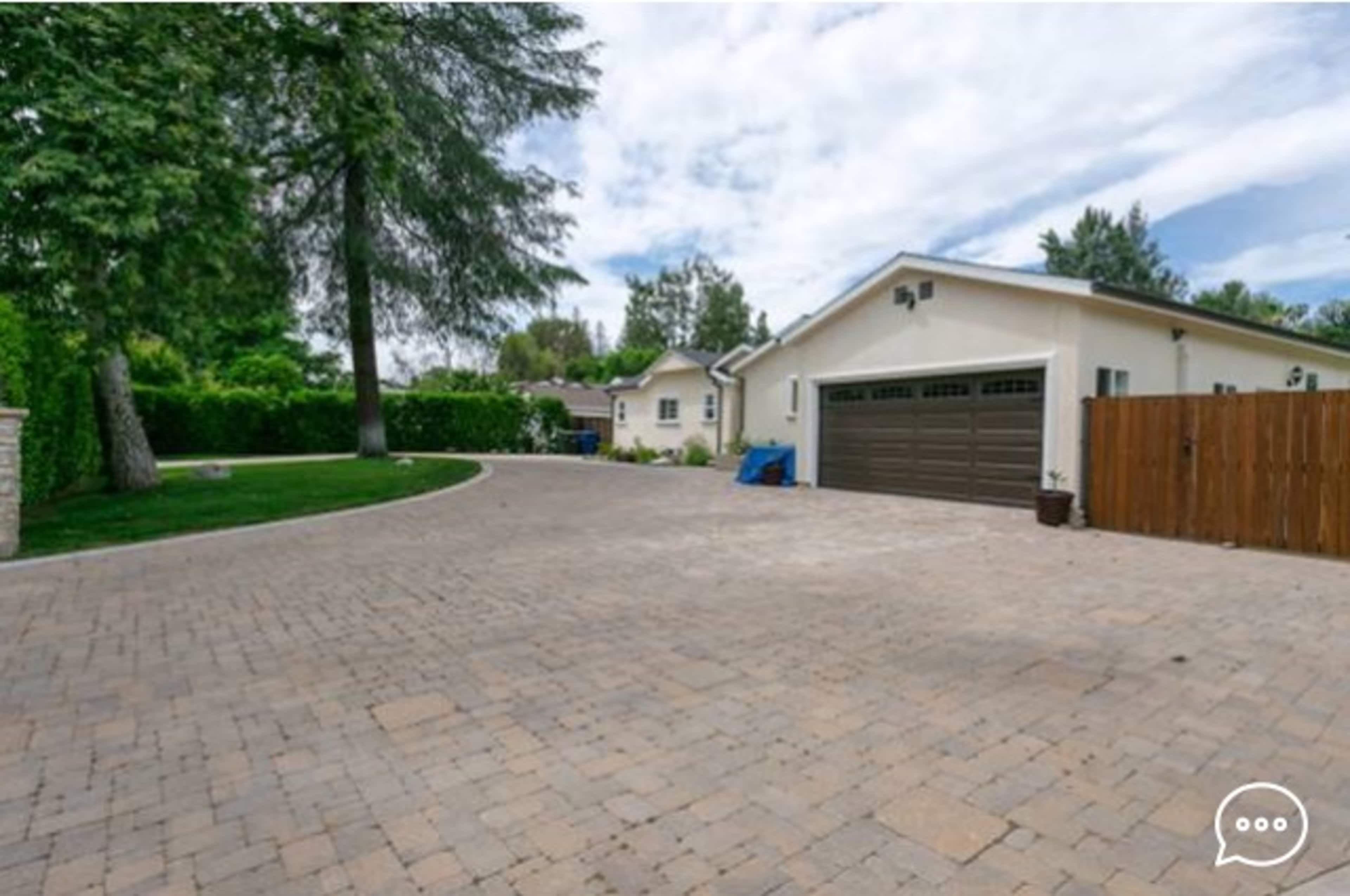 A paved driveway leads to a garage and a house, surrounded by greenery and trees.