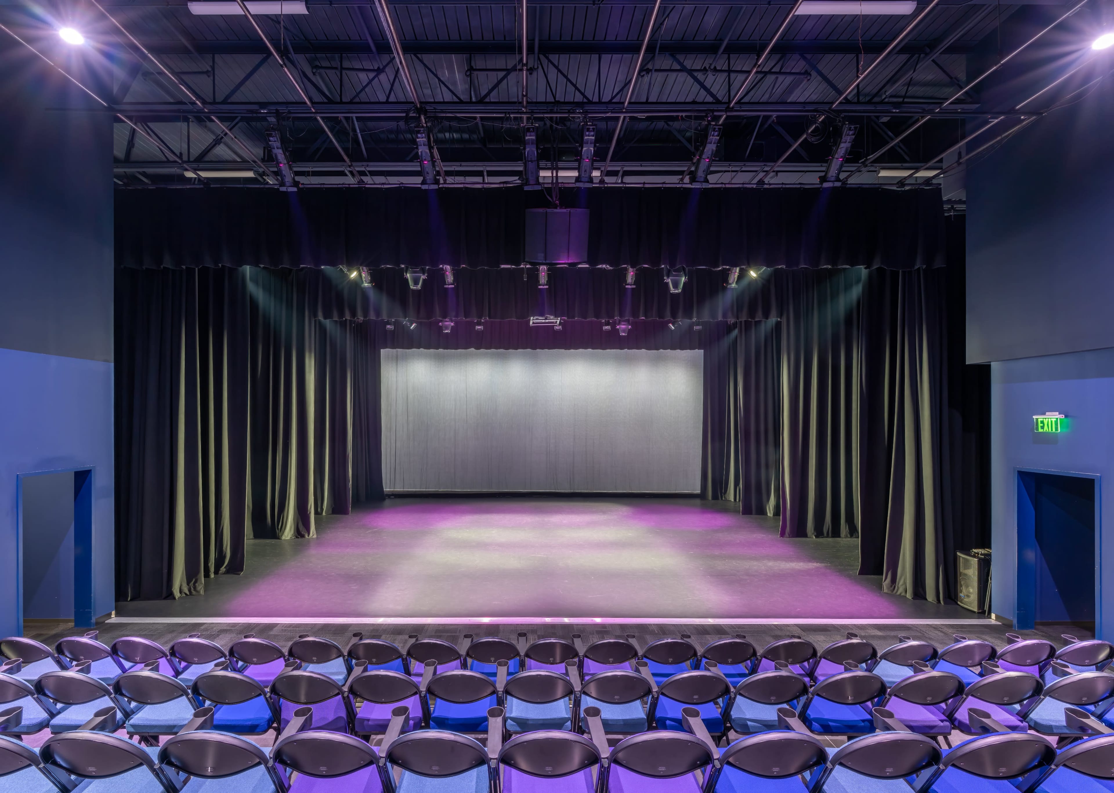 The image shows a theater stage with a dark curtain backdrop and rows of empty blue chairs facing the stage.
