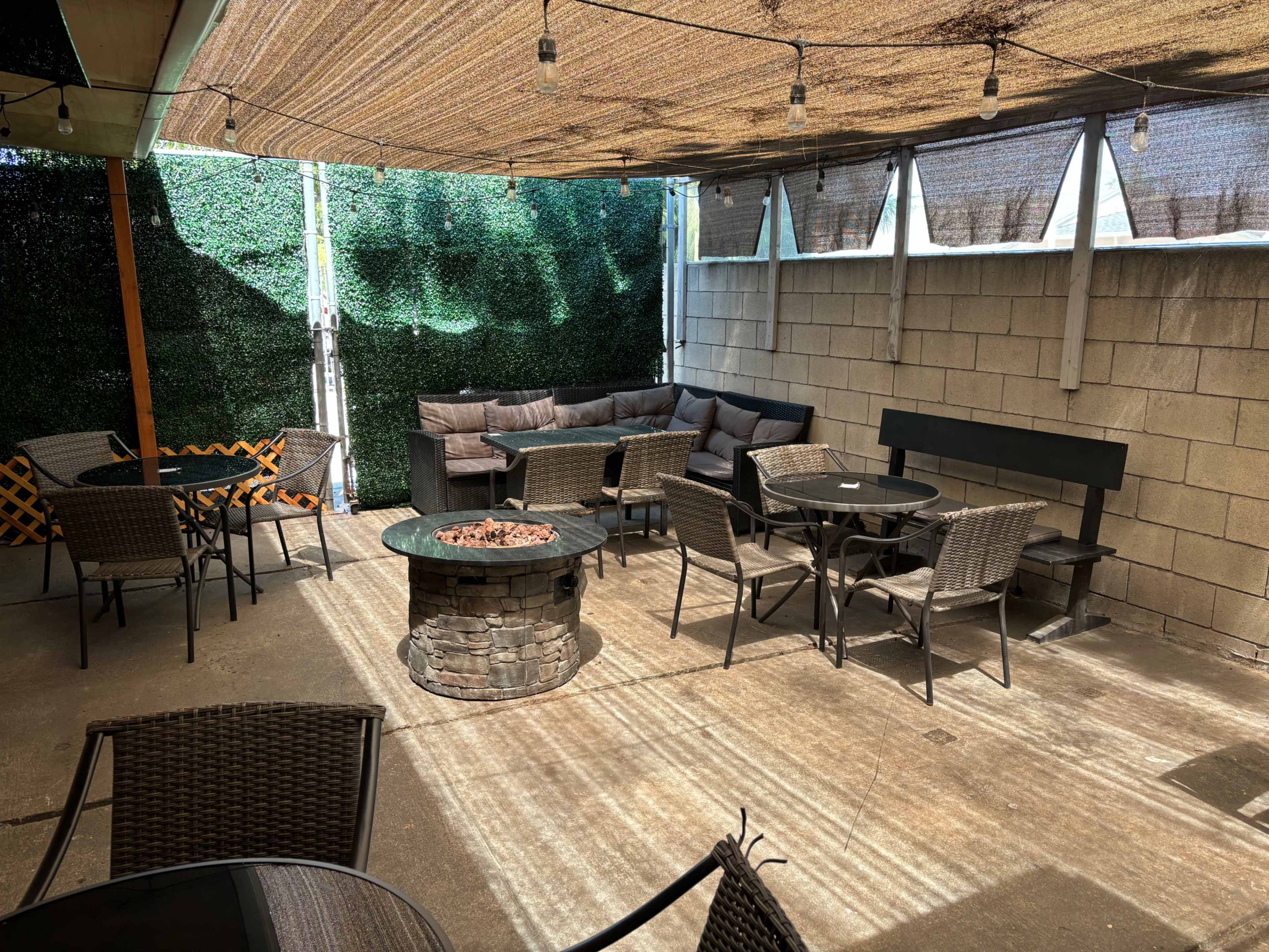 A shaded outdoor patio area with several tables, chairs, and a seating arrangement around a circular stone fire pit.