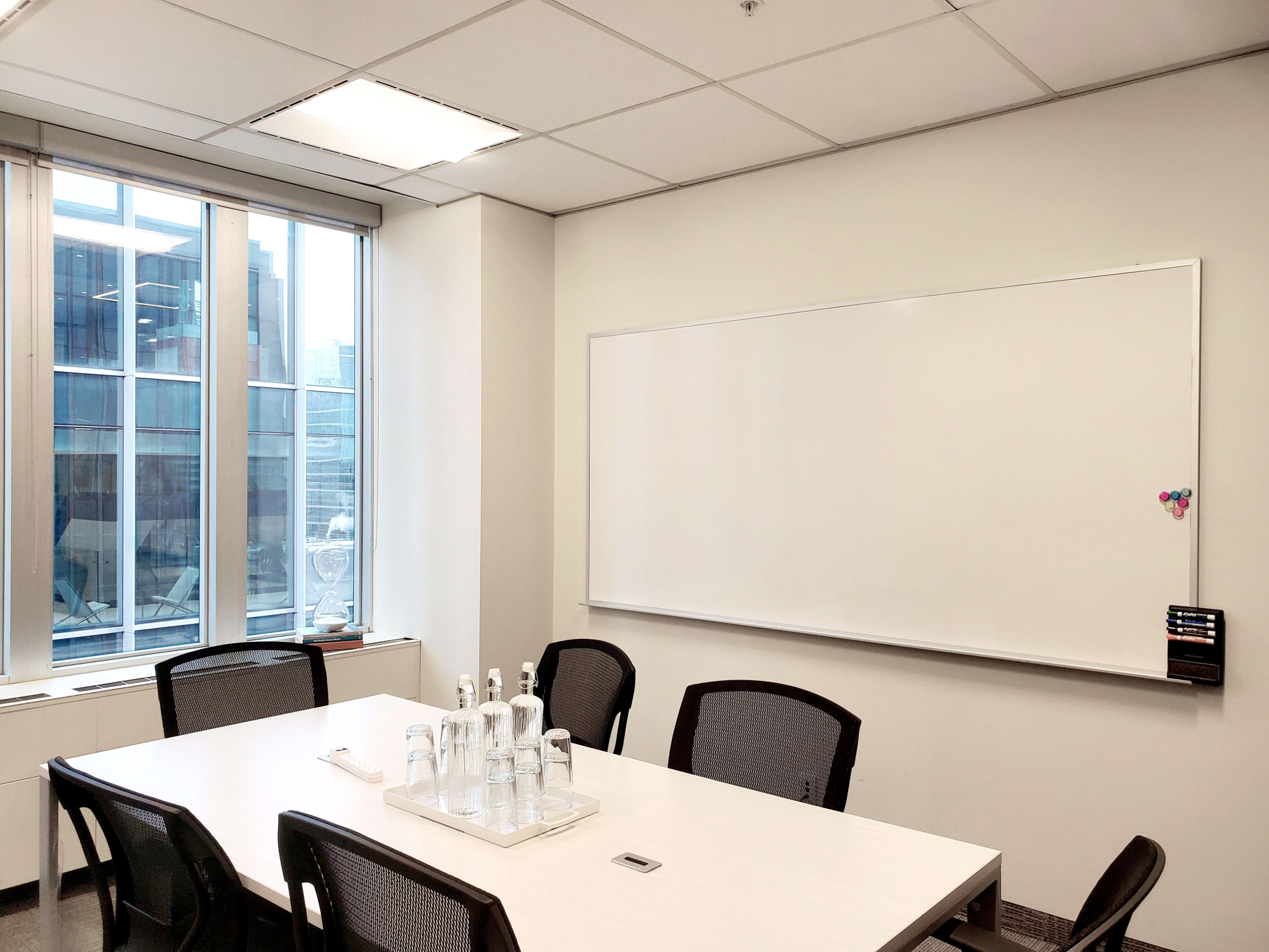 A modern conference room features a large whiteboard, a rectangular table with chairs, and natural light streaming in from large windows.