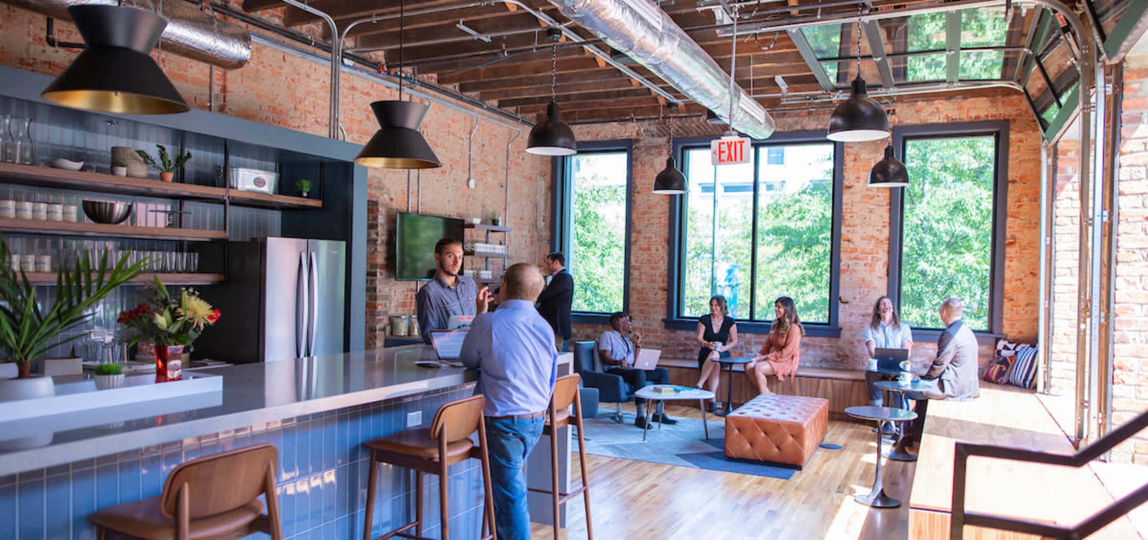 The image shows a modern co-working space with a kitchen area featuring a long bar, several people engaged in conversation, and large windows allowing natural light to fill the room.