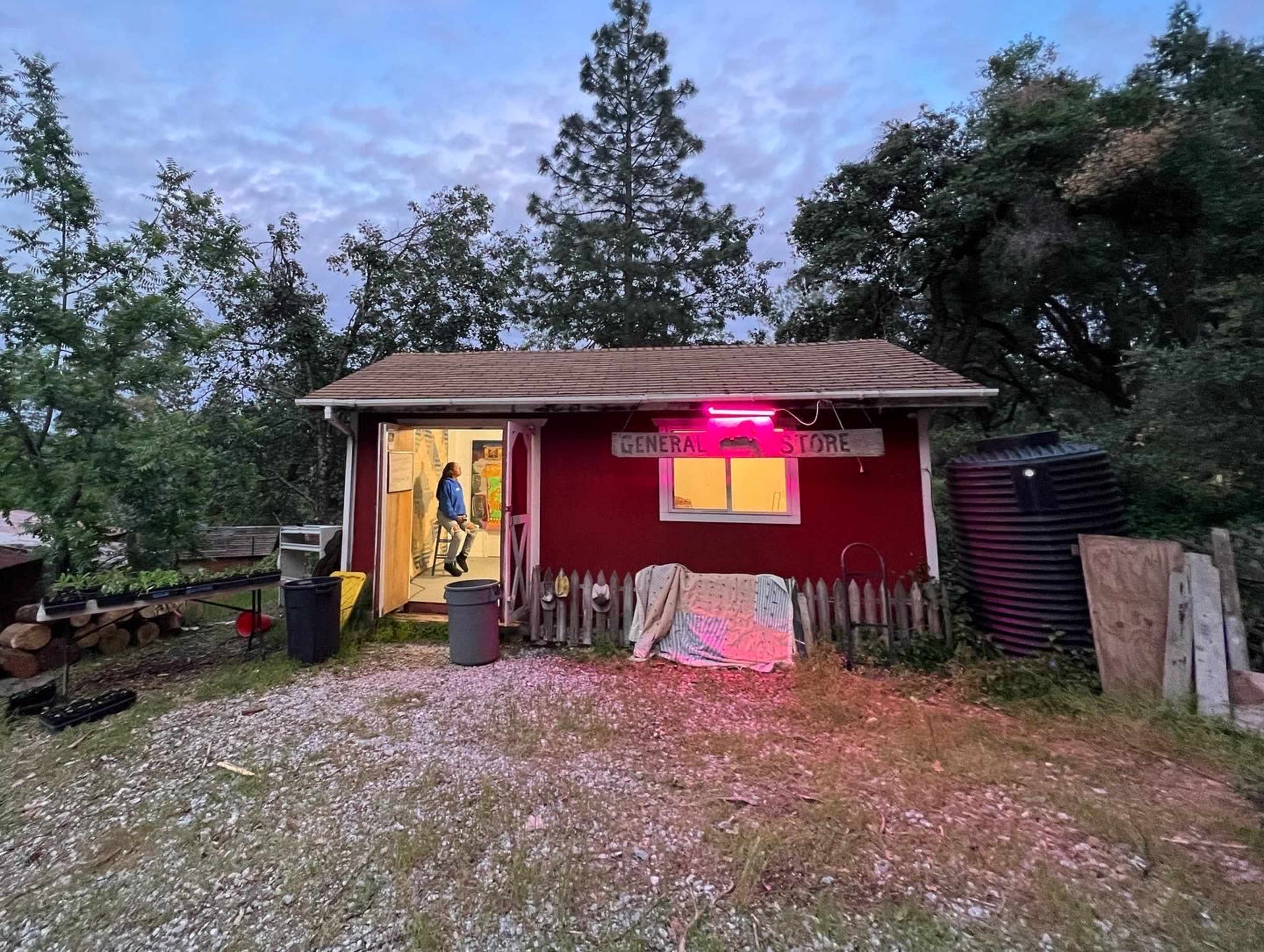 A red general store with a sign hangs above its entrance, where a figure stands inside by the open door, surrounded by trees and gravel.