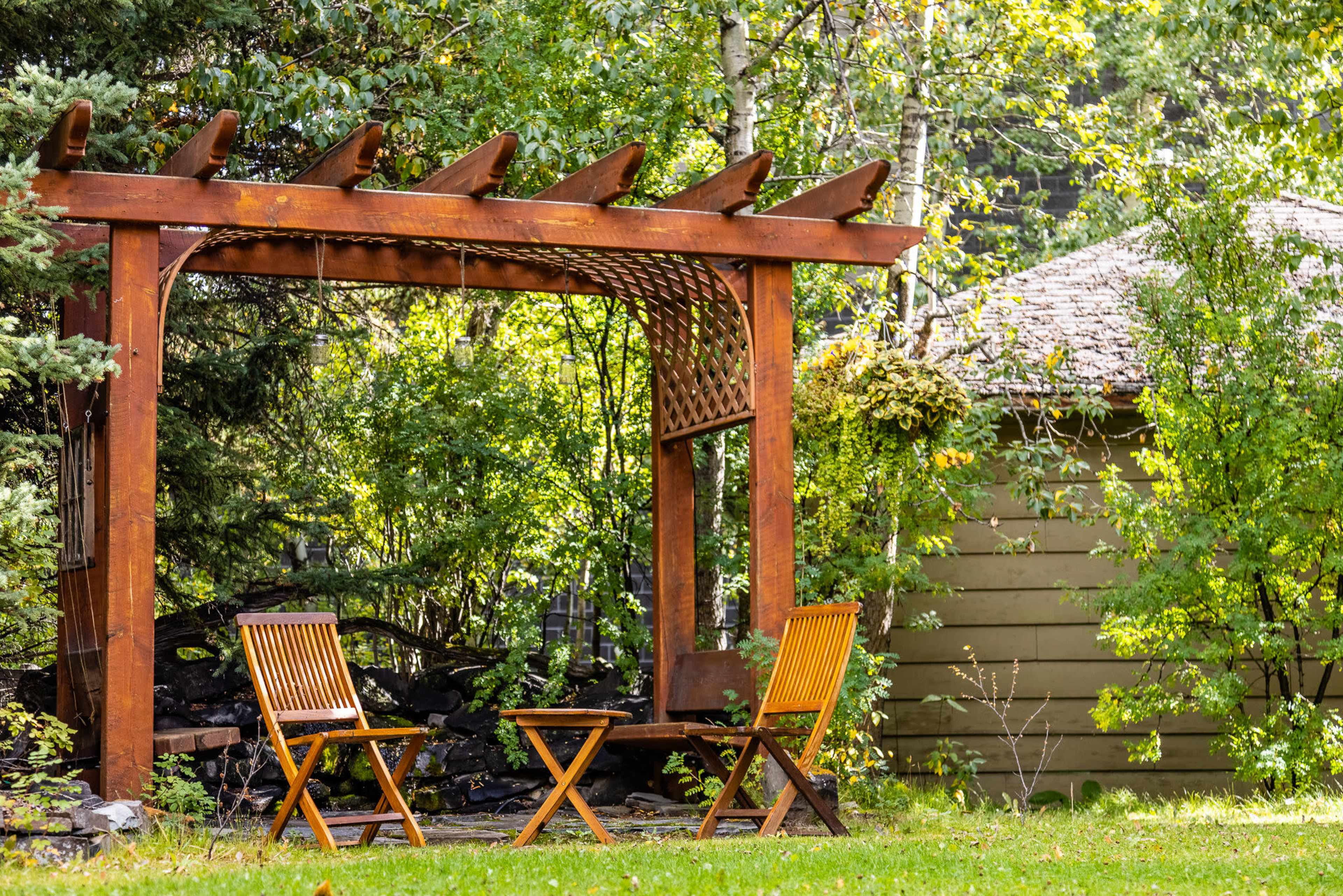 A wooden pergola with two folding chairs is set in a garden surrounded by trees and a rustic shed in the background.