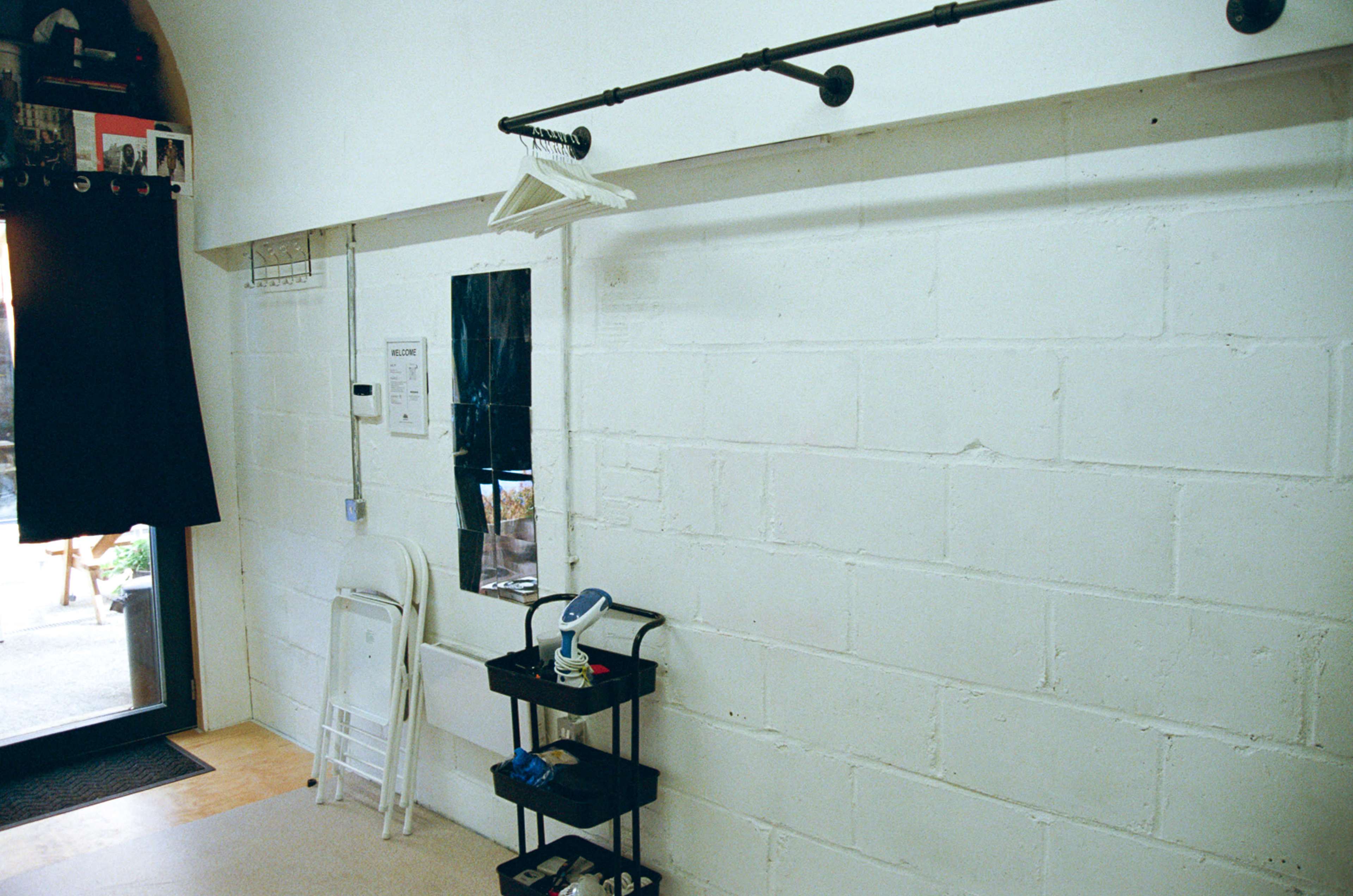 The image shows a bare white wall with a coat rack, a mirror, a folding chair, and a small cart with various items.