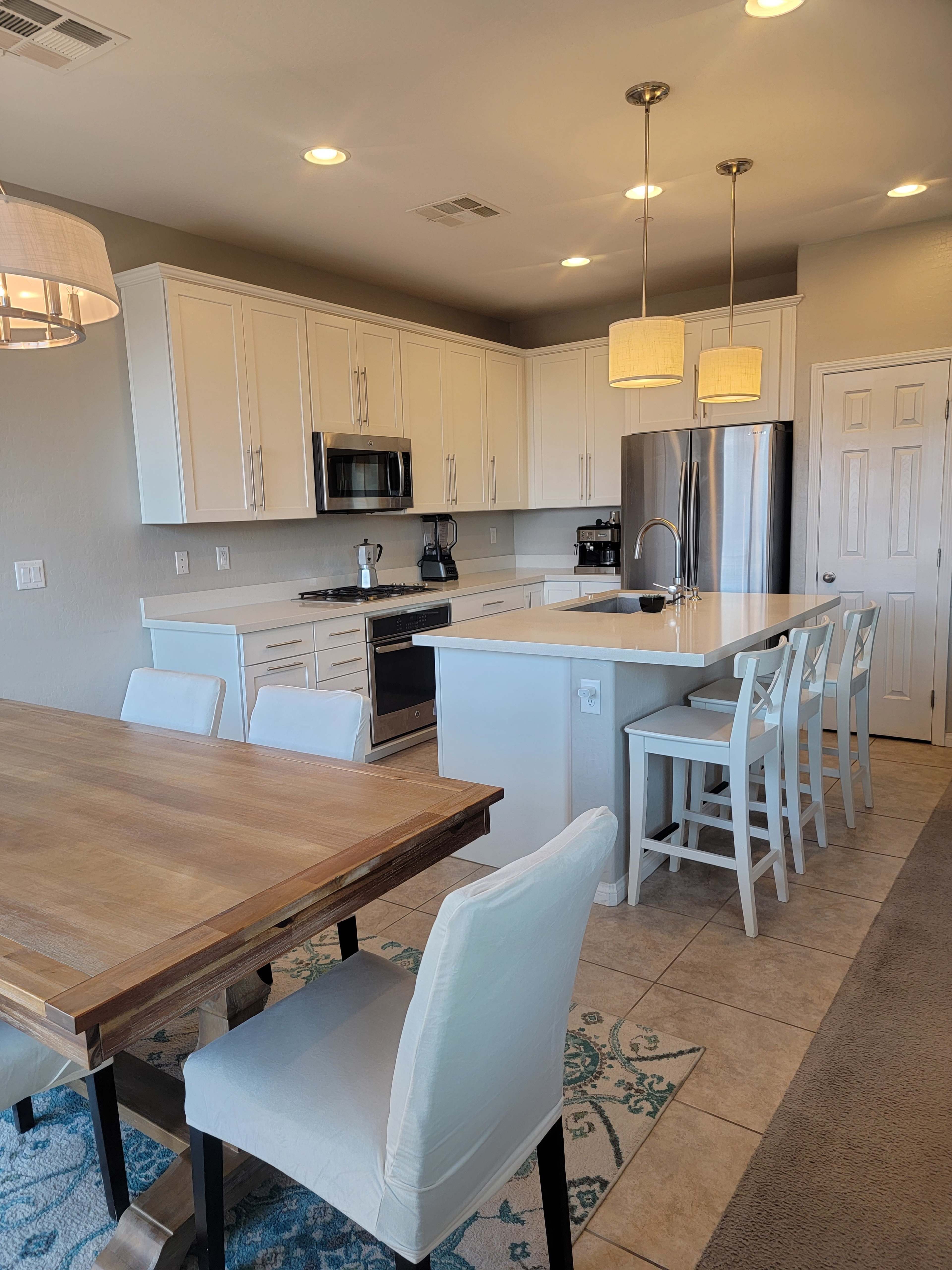 A modern kitchen features white cabinets, stainless steel appliances, and a dining area with a wooden table and white chairs.